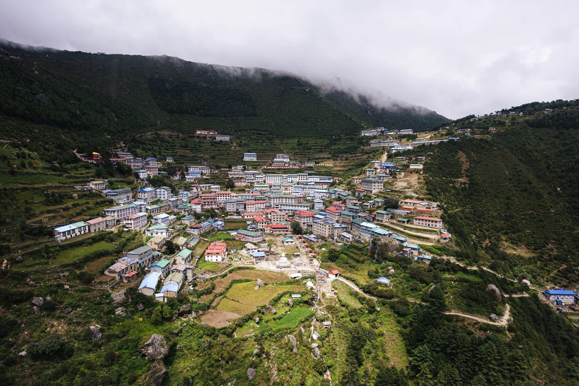 Aerial view of Namche Bazaar, the gateway town to the Mt Everest region, clinging to a steep Khumbu hillside under low cloud.