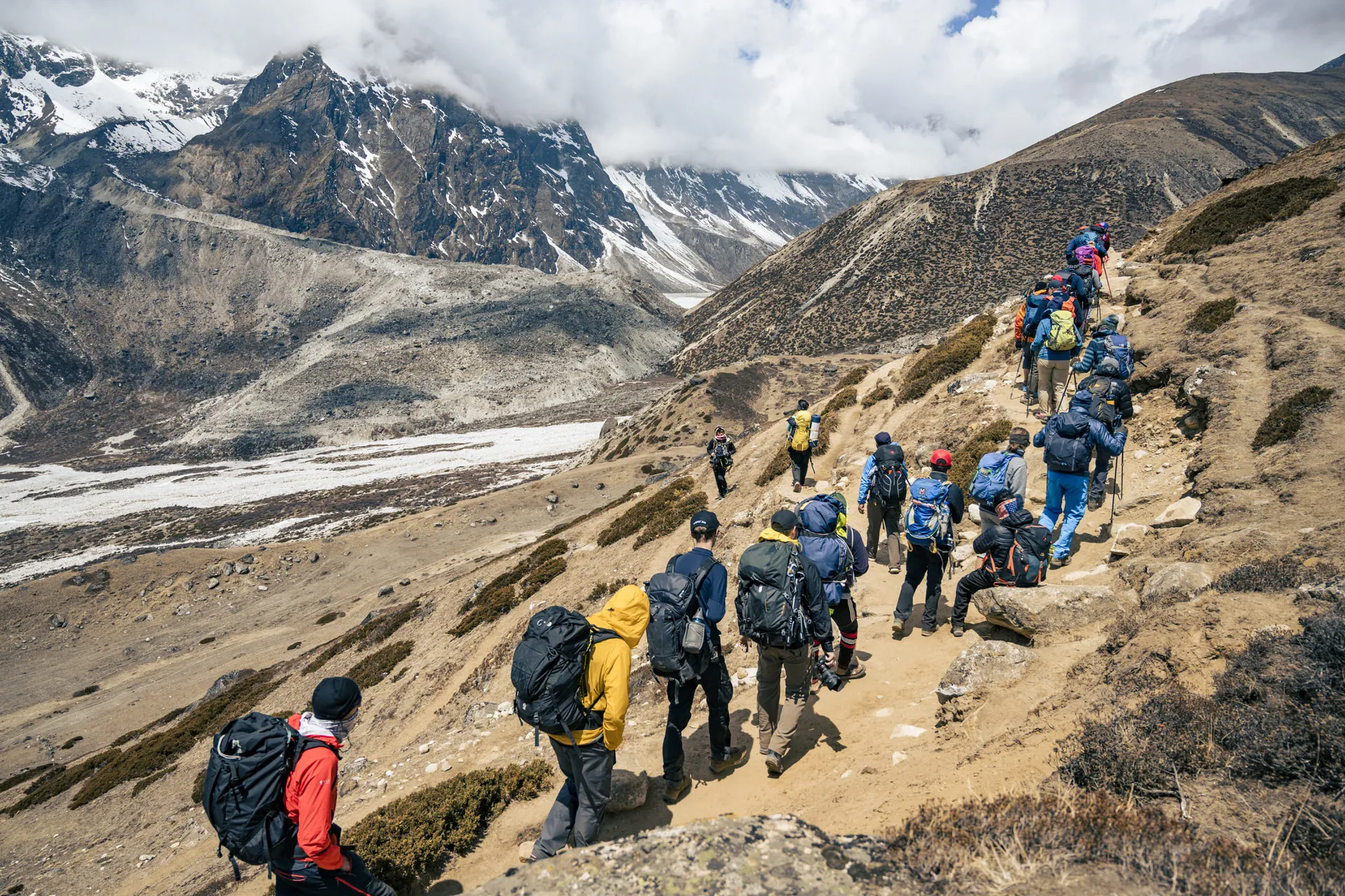 A long line of trekkers walks single-file along a sandy trail high on the Khumbu plateau, snow peaks rising ahead.