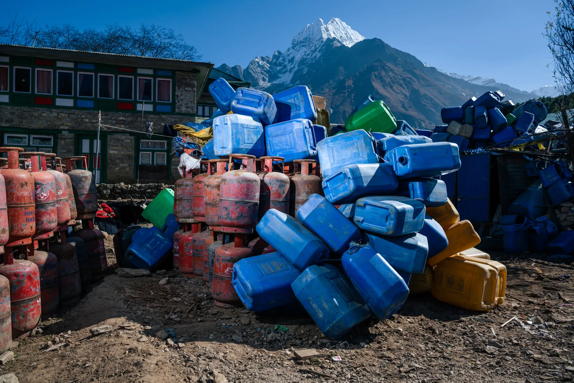 Empty blue and yellow plastic jerry cans and red gas cylinders piled in a Khumbu yard, with mountains rising behind the village.