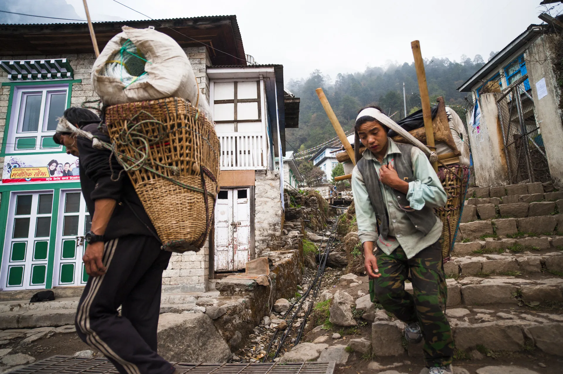 Two Sherpa porters carry tall bamboo baskets and long wooden poles up steep stone steps in a Khumbu village.