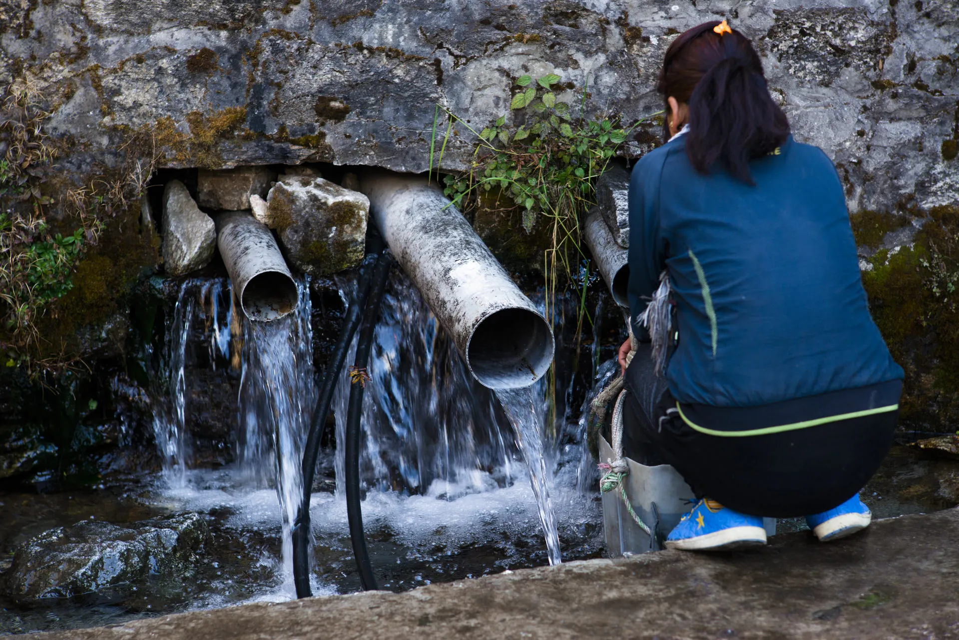 A young Nepali woman crouches at a public concrete water tap to fill a container from pipes set into a stone wall.