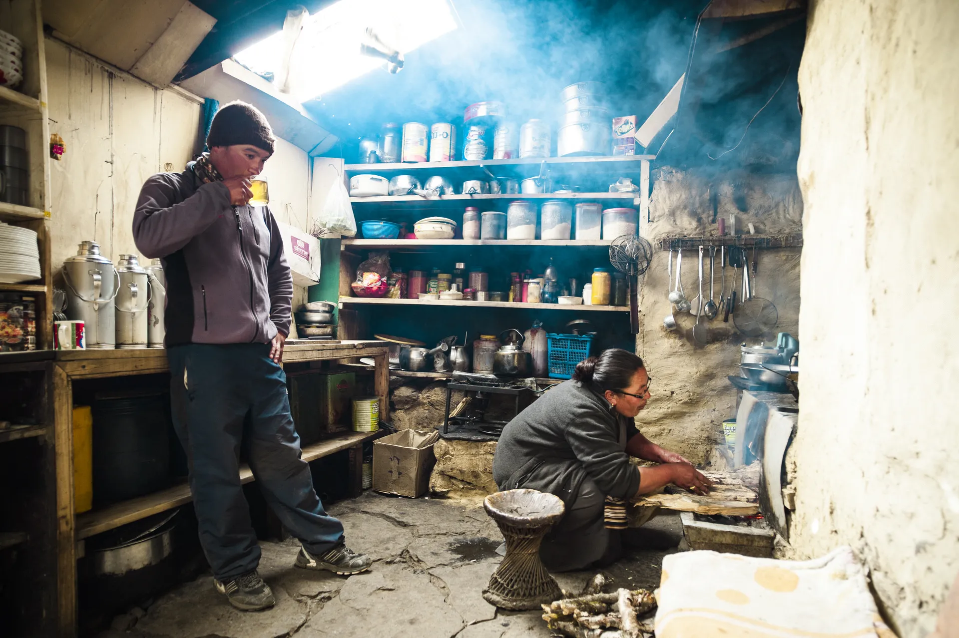 Two Sherpas in a smoky Khumbu kitchen — one standing with a yellow mug, the other crouched at a wood stove preparing food.