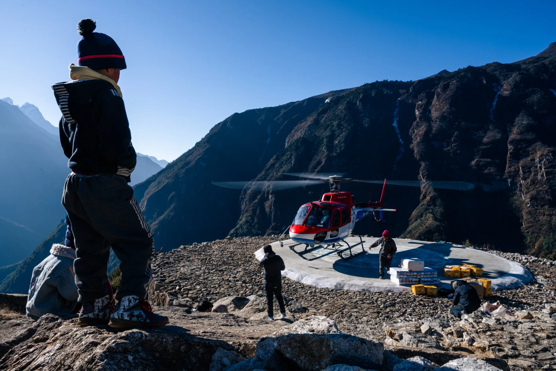 A red supply helicopter on a stone helipad above Namche Bazaar, men in jackets watching as orange crates are unloaded.