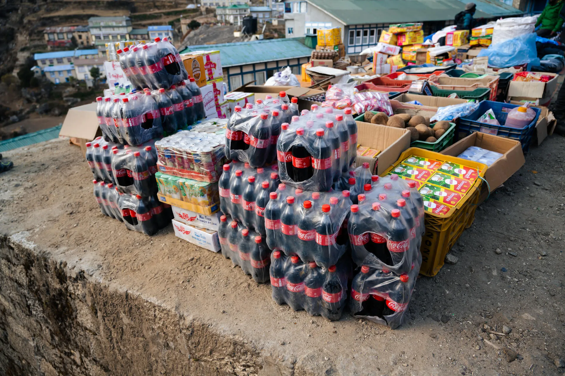 Cases of plastic-wrapped Coca-Cola, bottled water and other drinks stacked beside boxes of fruit on a Namche shop wall.