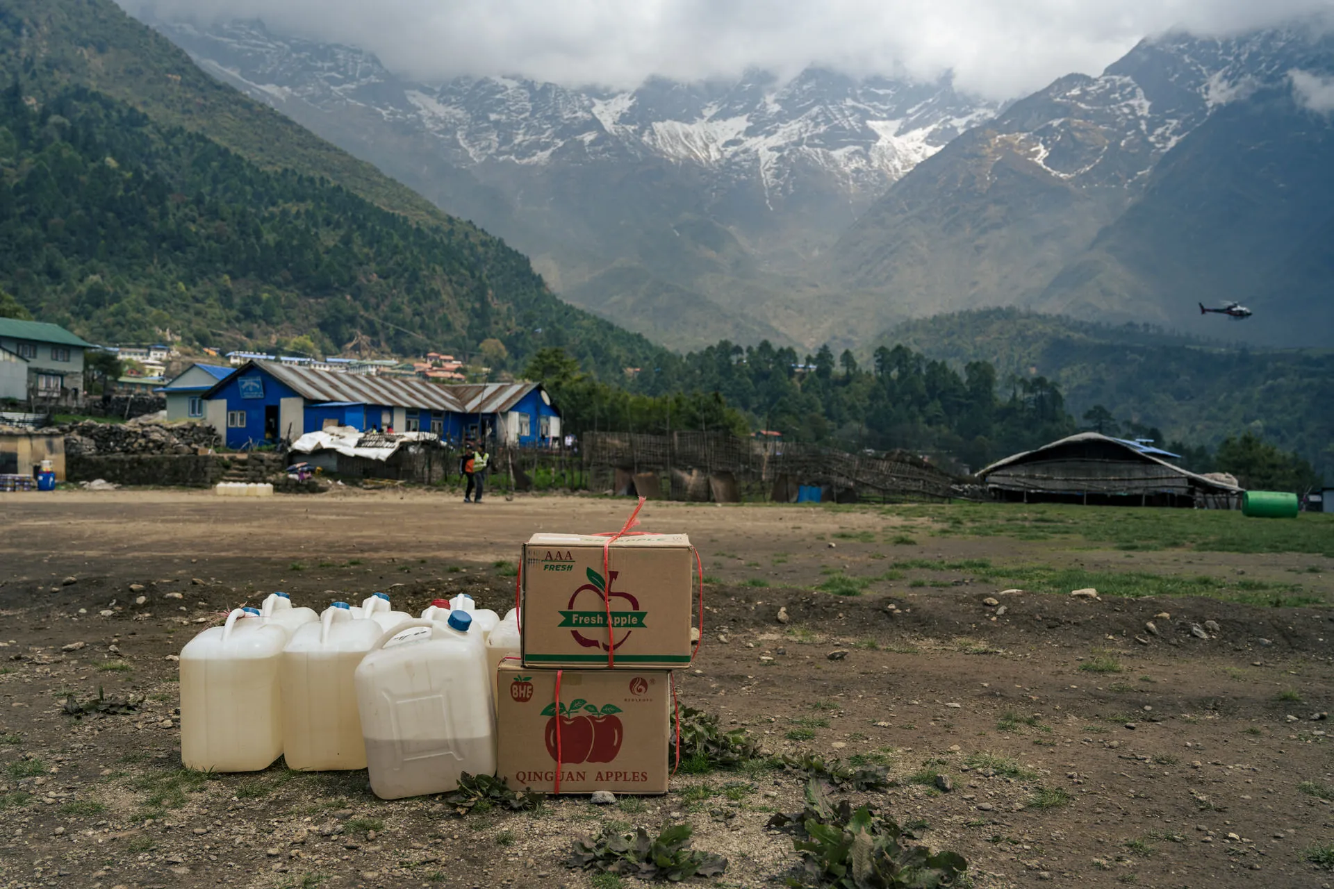 White plastic jerry cans of fuel and cardboard boxes of imported apples wait beside the Lukla airstrip, a helicopter approaching across the mountains.