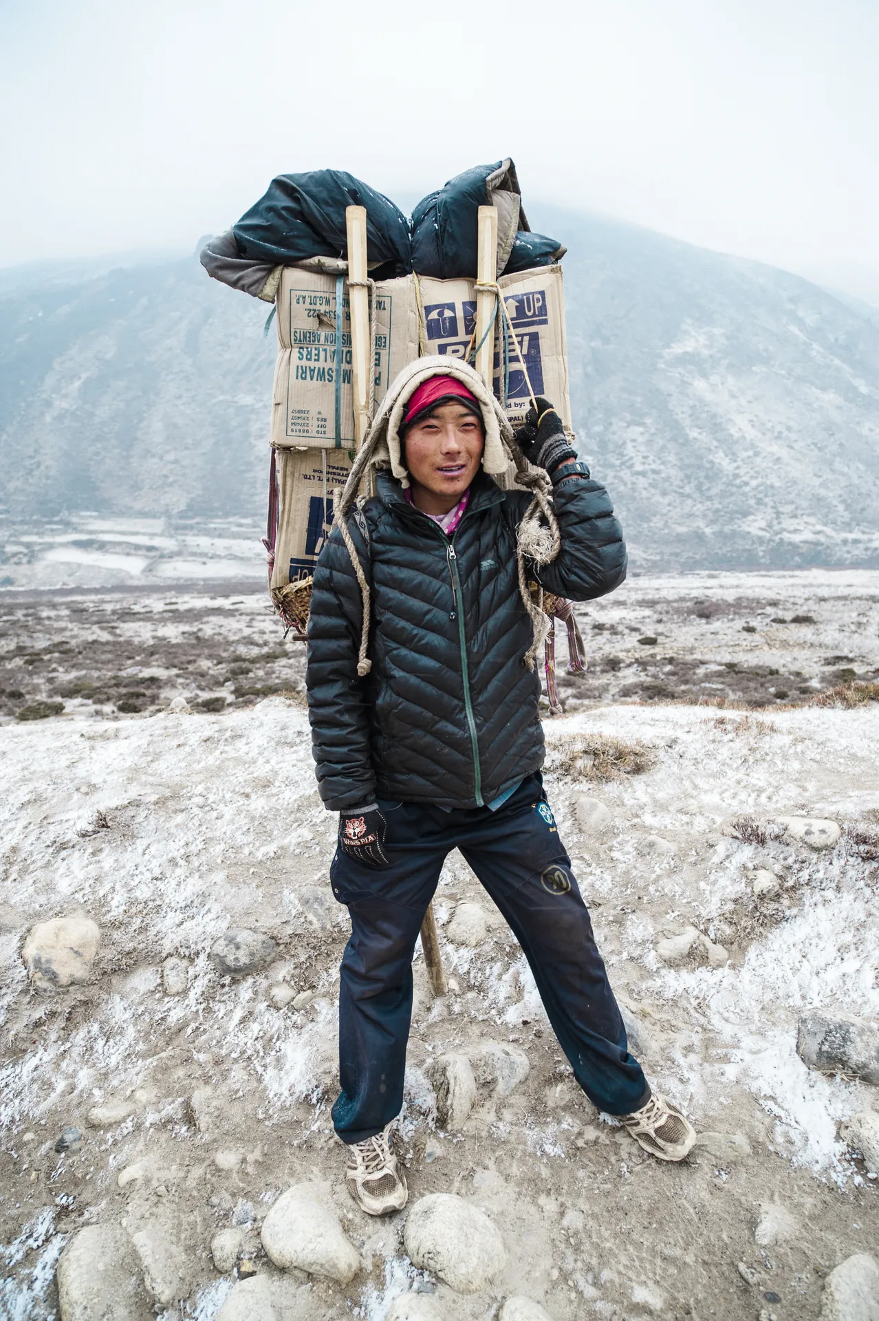 A young Sherpa porter stands on the trail with an enormous load of cardboard supplies on his back, supported by a tumpline across his forehead.