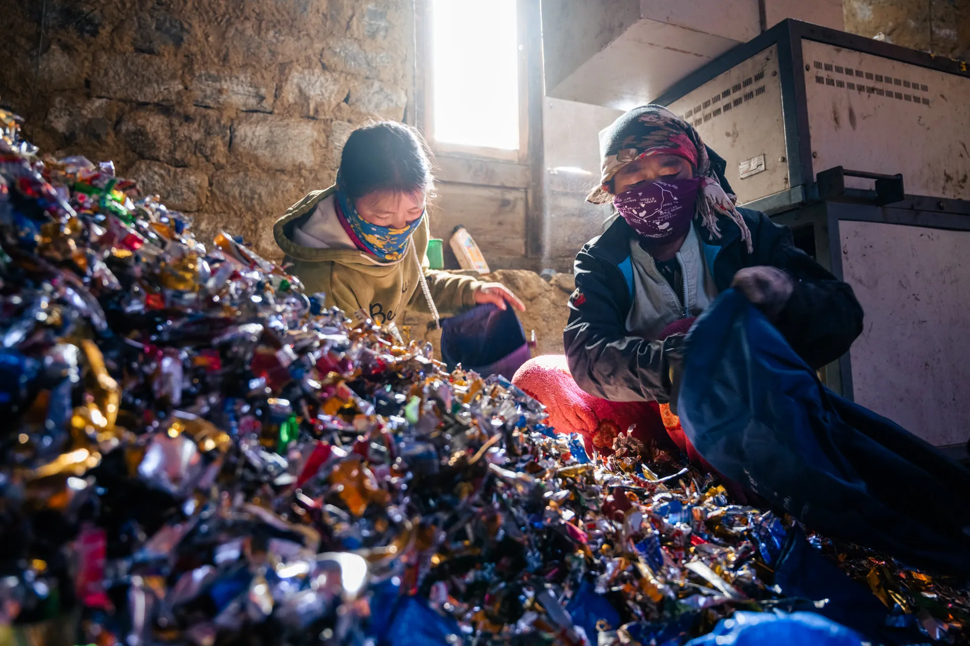 Two waste workers in masks sort heaps of crushed foil packets and snack wrappers at a Sagarmatha Pollution Control Committee facility.