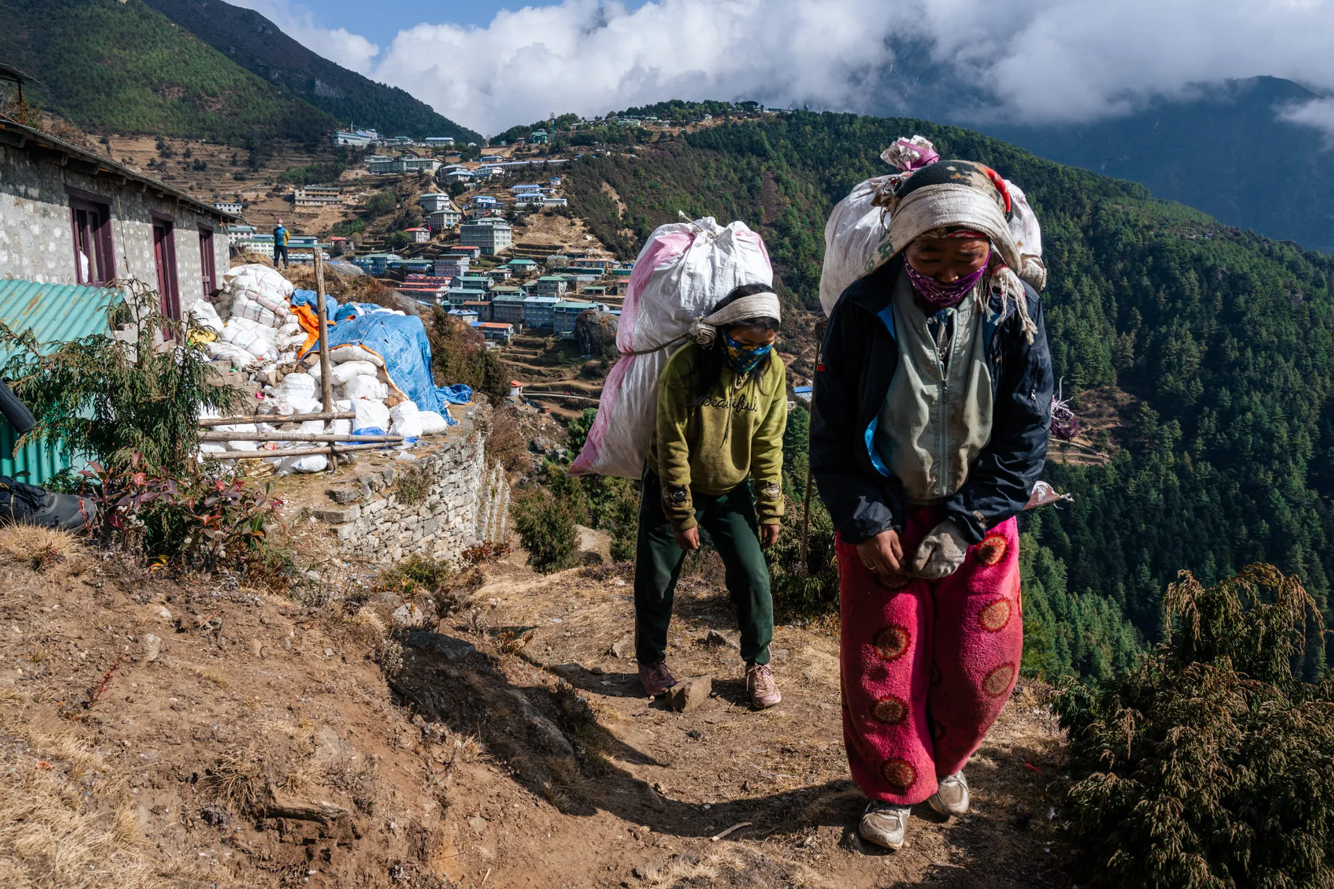Two Sherpa women carry large sacks of household waste along a path above Namche Bazaar, masks pulled over their faces.