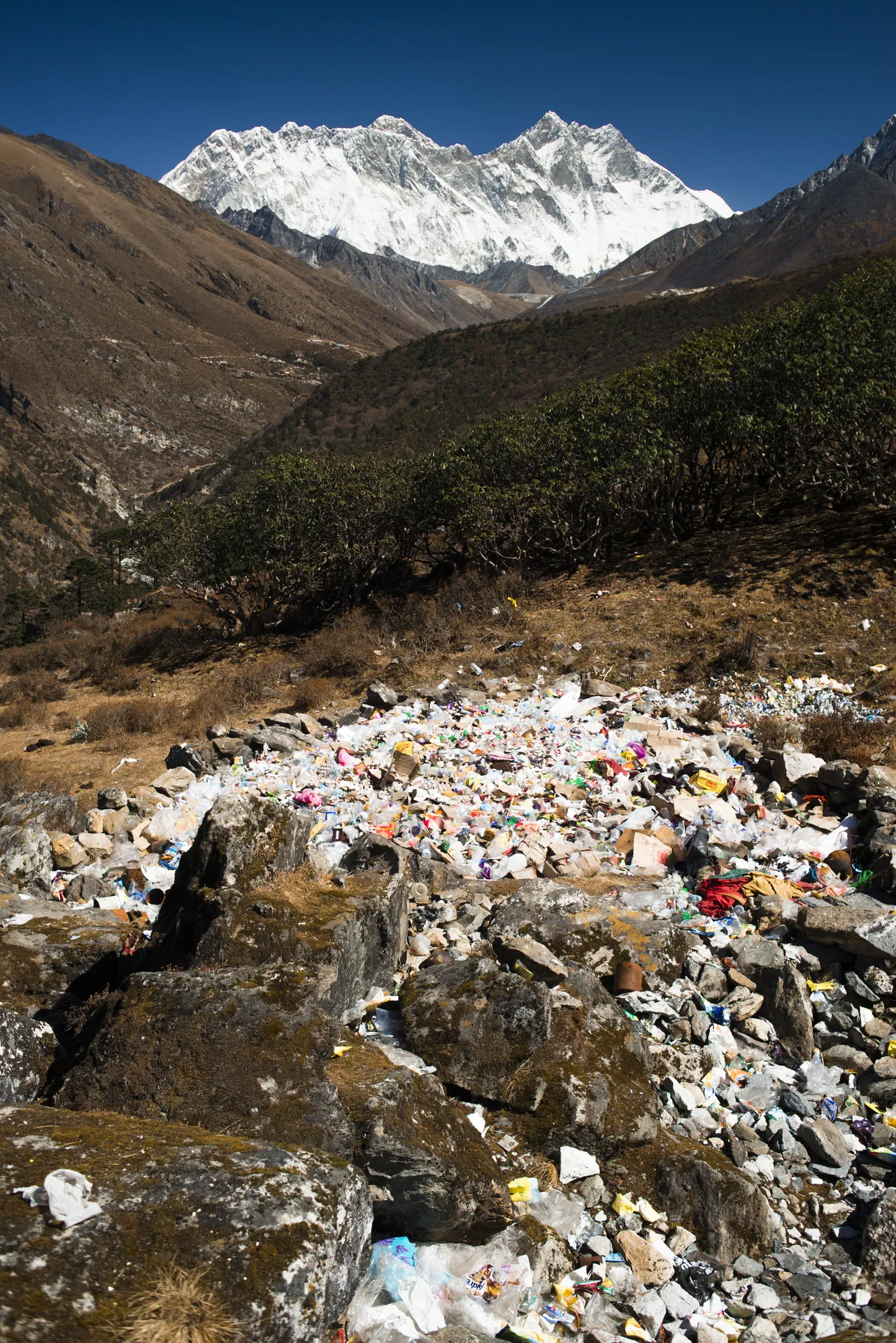 A pile of plastic bottles and packaging litters the rocks of an alpine creek with the snow face of Lhotse rising behind.