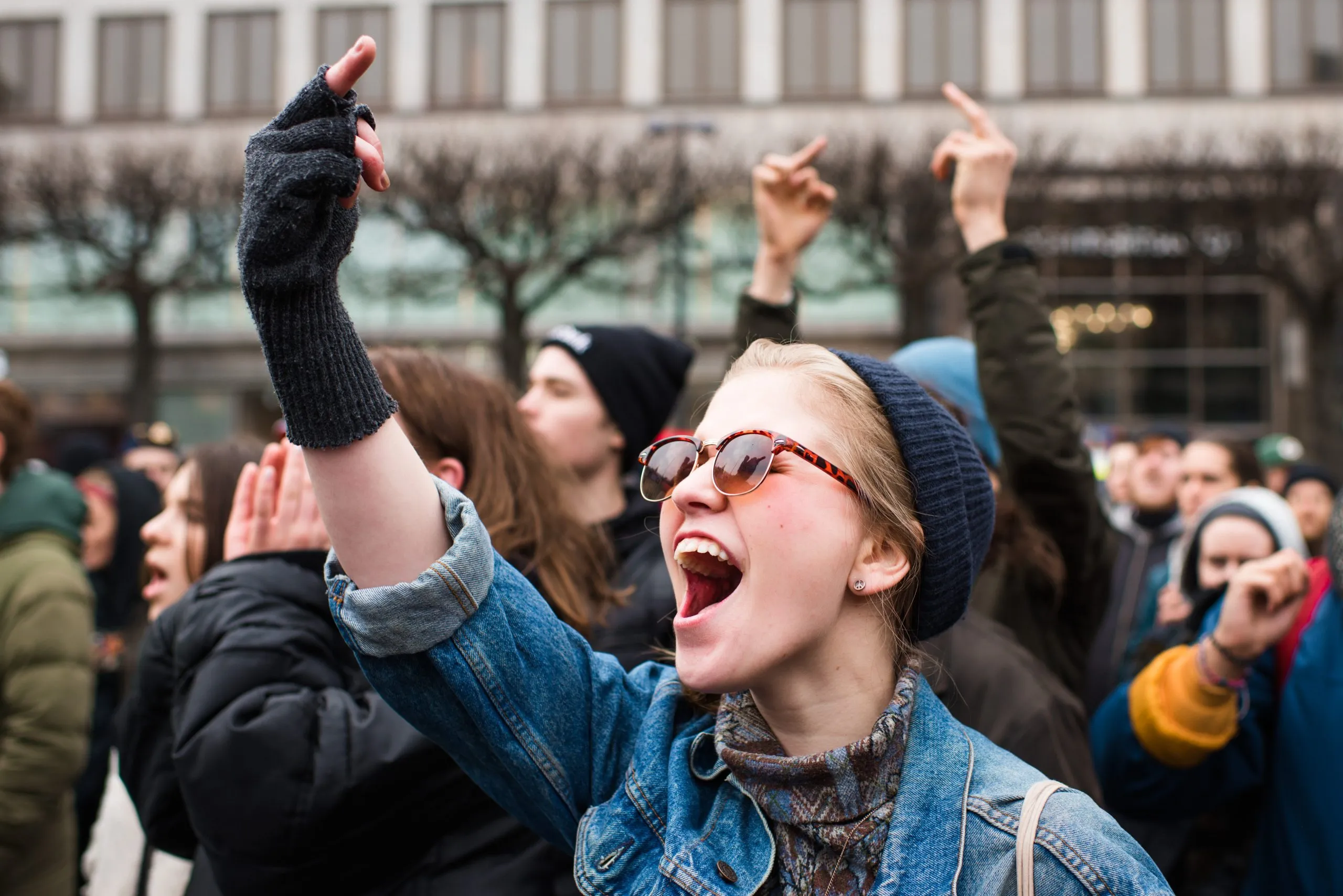 A young woman in a denim jacket, beanie and sunglasses cheers and raises a fingerless-gloved hand at a daytime rally in Stockholm.