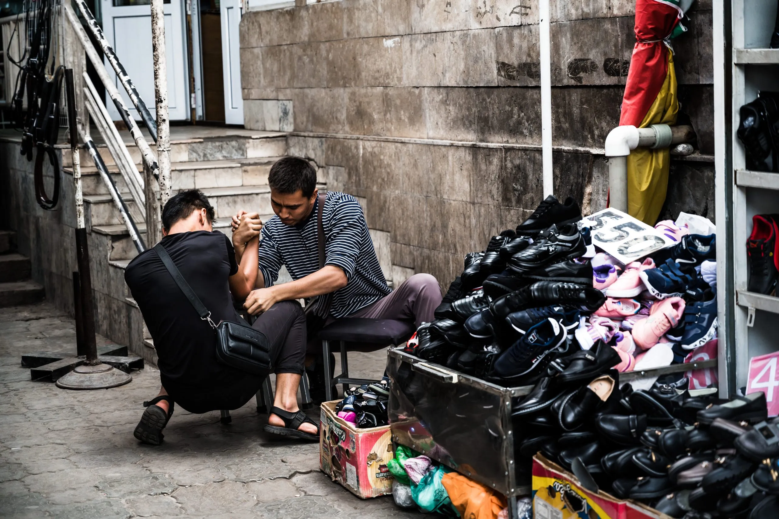 Two men arm-wrestle on a small stool beside a market shoe stall on a street in Bishkek, Kyrgyzstan.