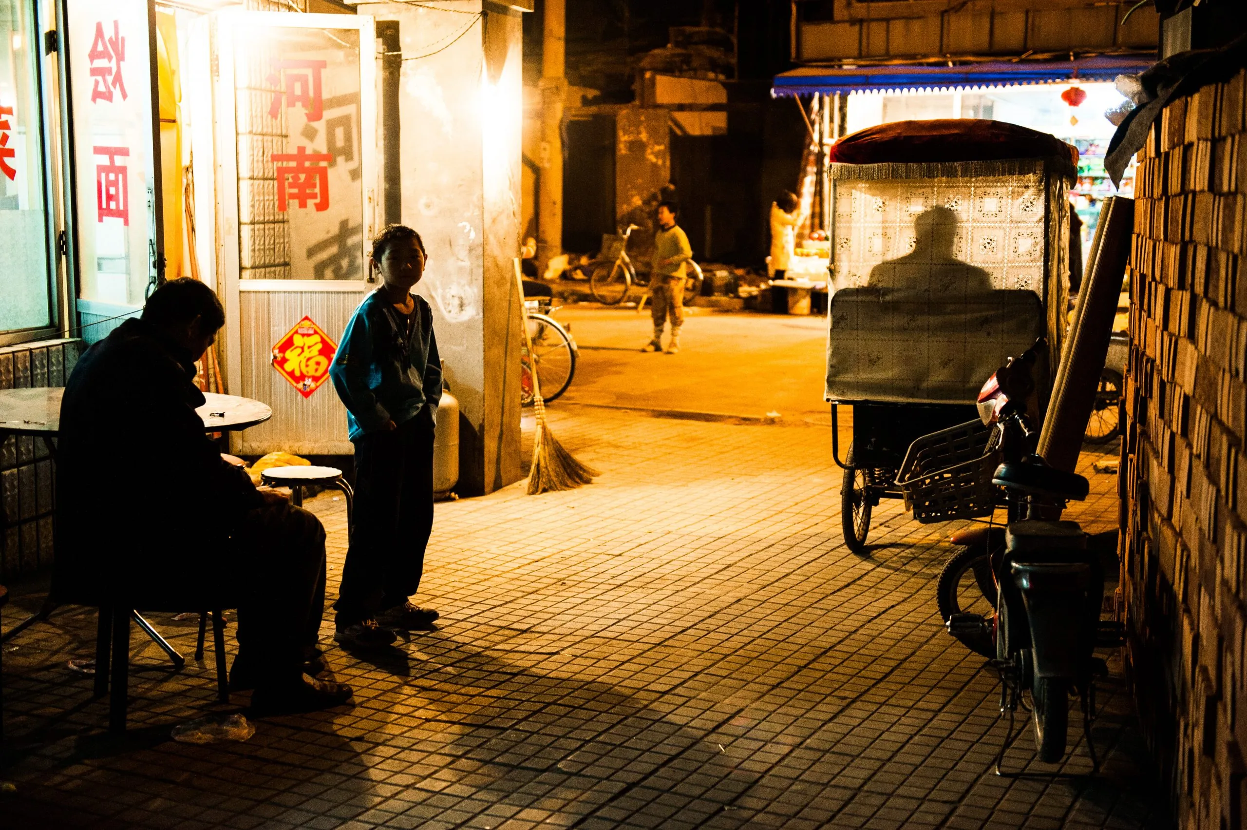 A Chinese woman stands beside a small noodle shop on a quiet warmly-lit street at night, a parked pedicab rickshaw to one side.