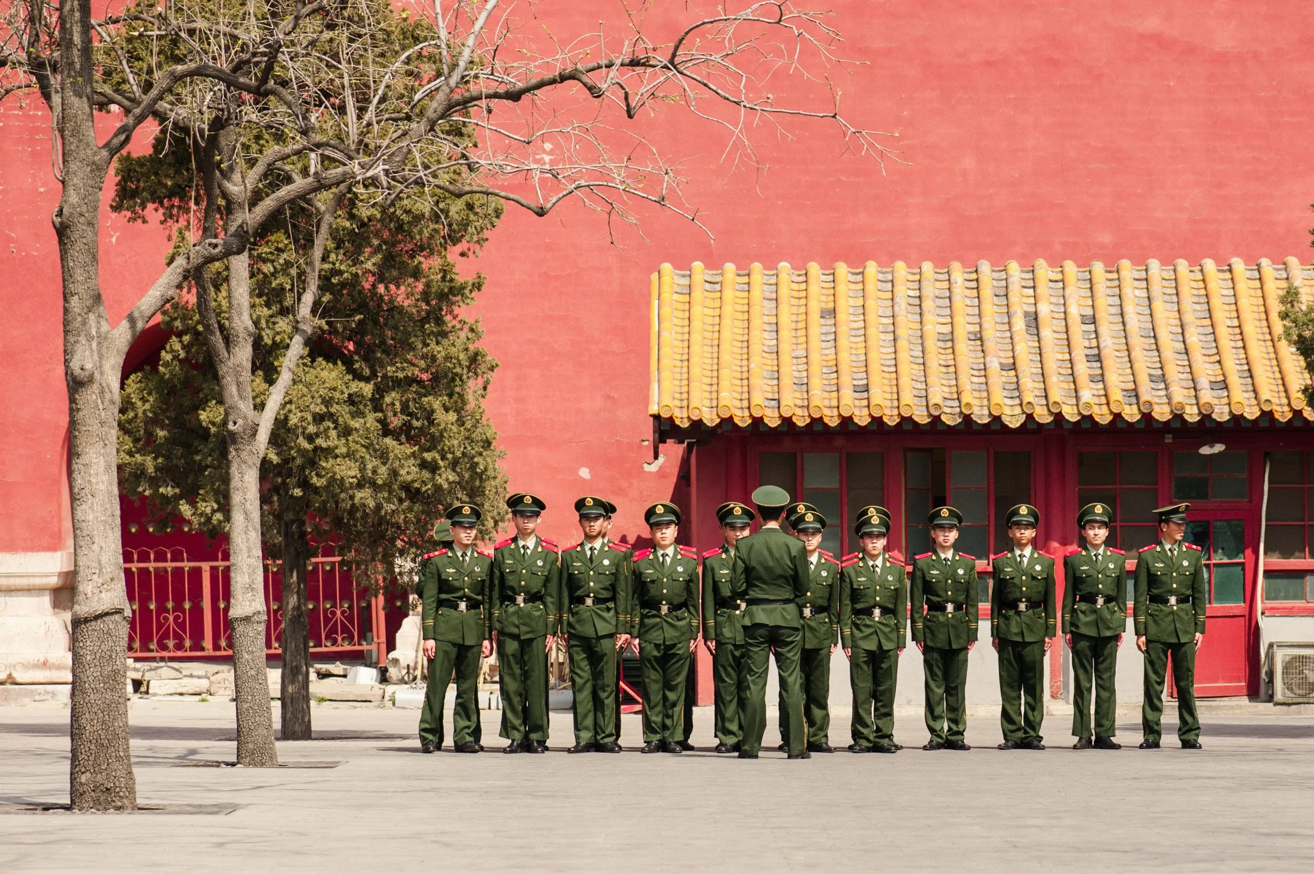 A line of Chinese paramilitary soldiers in green uniforms stand at attention in front of a red Forbidden City wall in Beijing.
