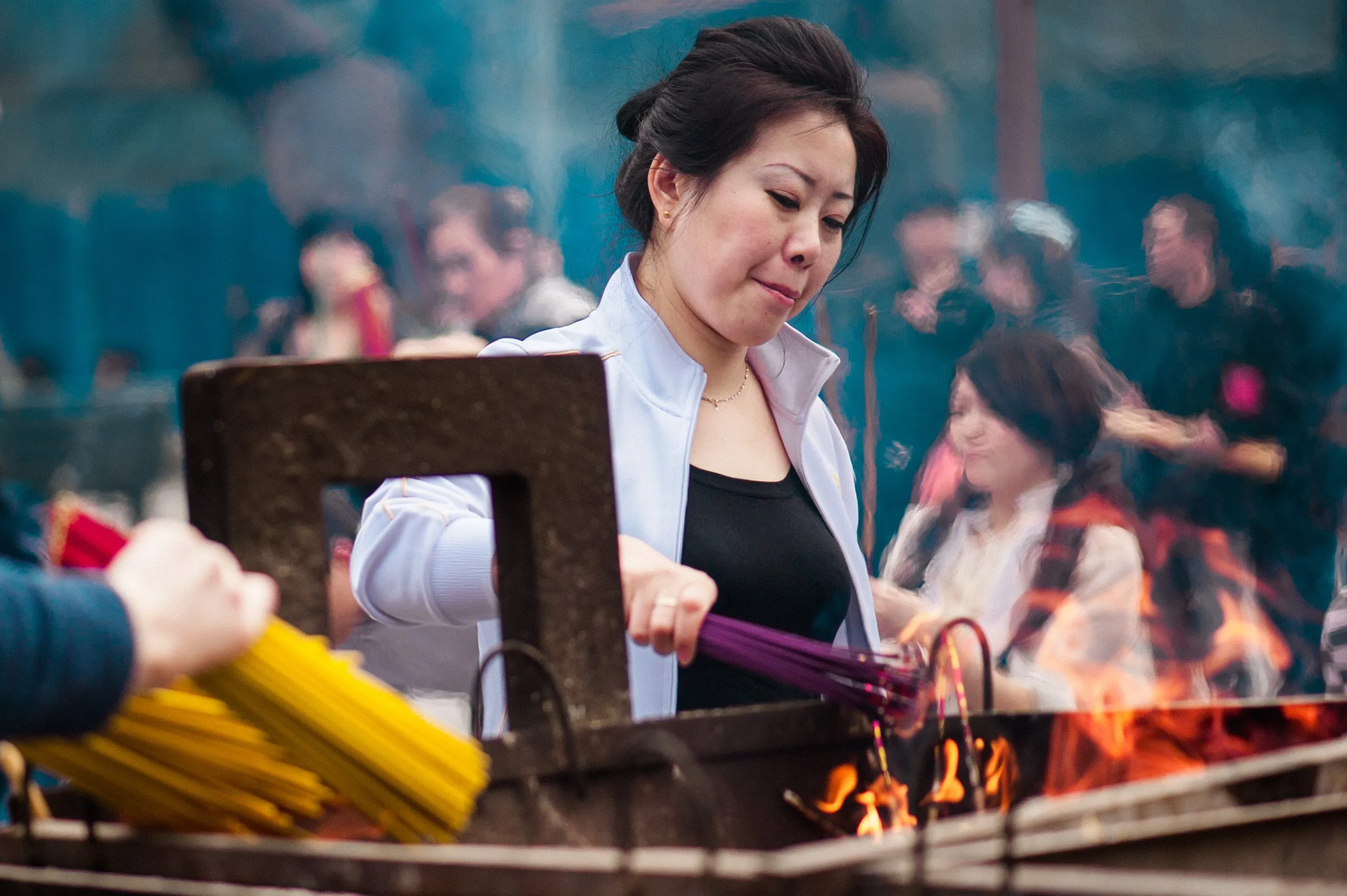 A woman lights a thick bundle of incense sticks at a Chinese Buddhist temple, smoke and other worshippers blurring around her.