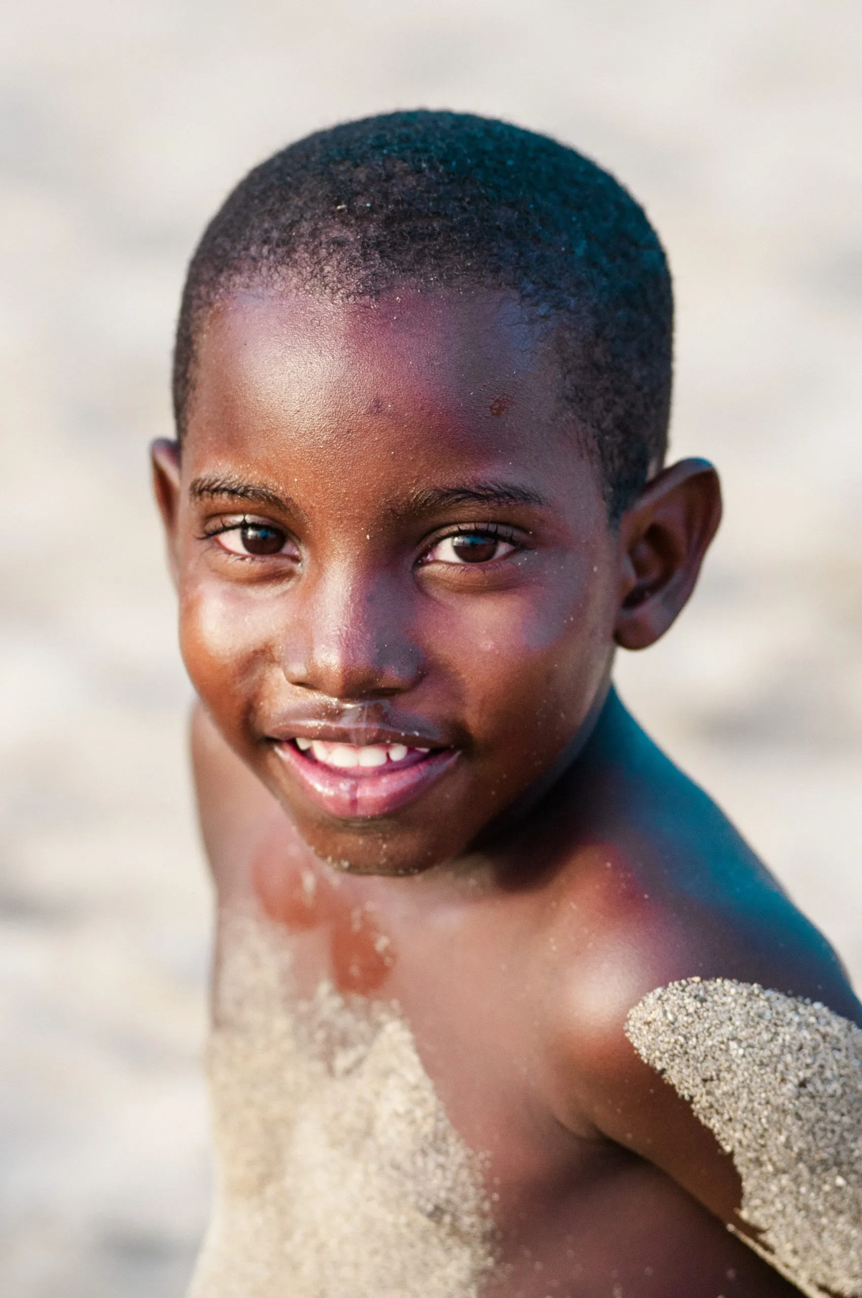 Close portrait of a Cape Verdean boy with sand on his shoulders smiling brightly on a sunlit beach.