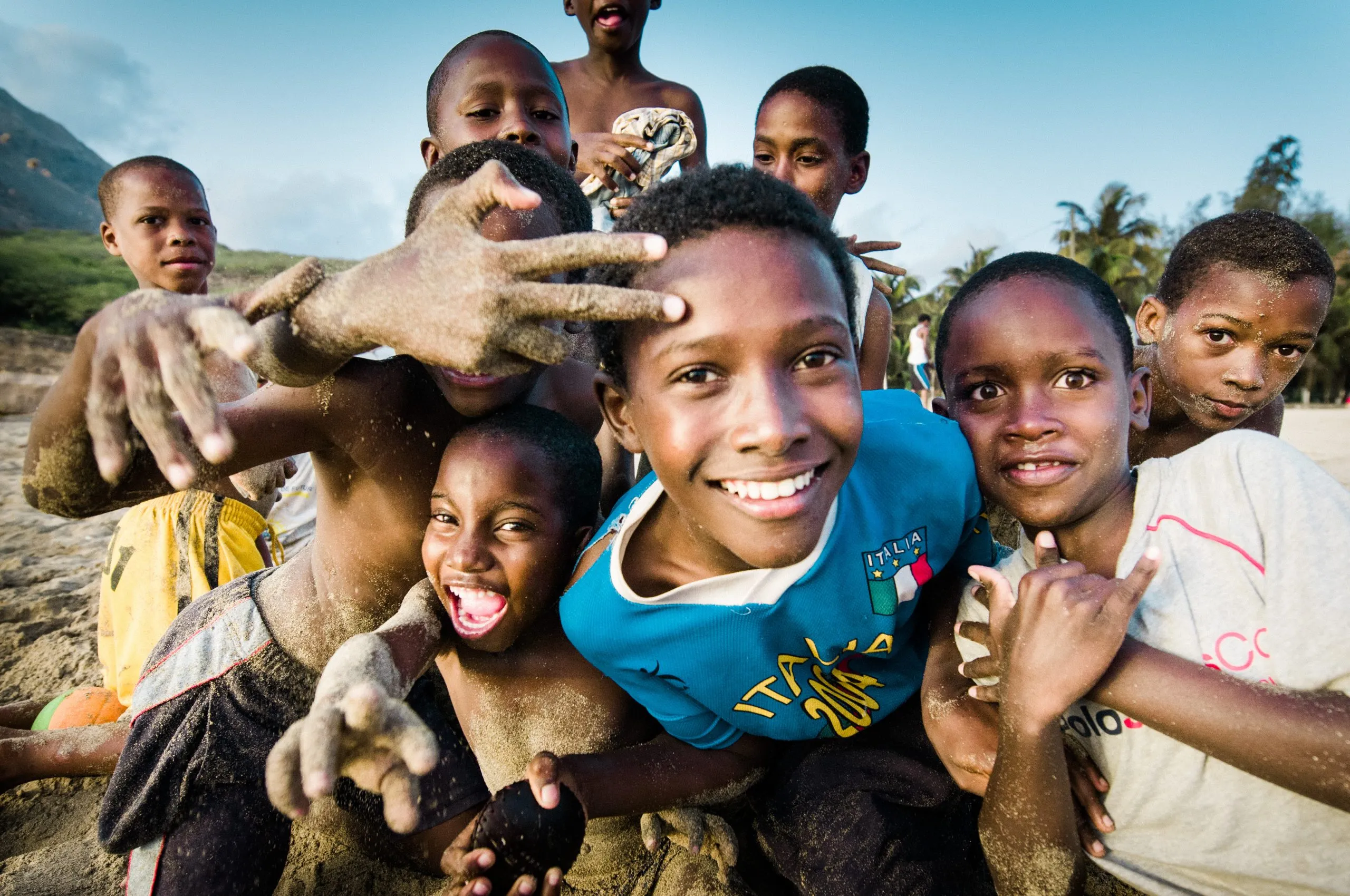 A group of Cape Verdean boys laugh and gesture toward the camera on a sandy beach, hands and shoulders dusted with sand.