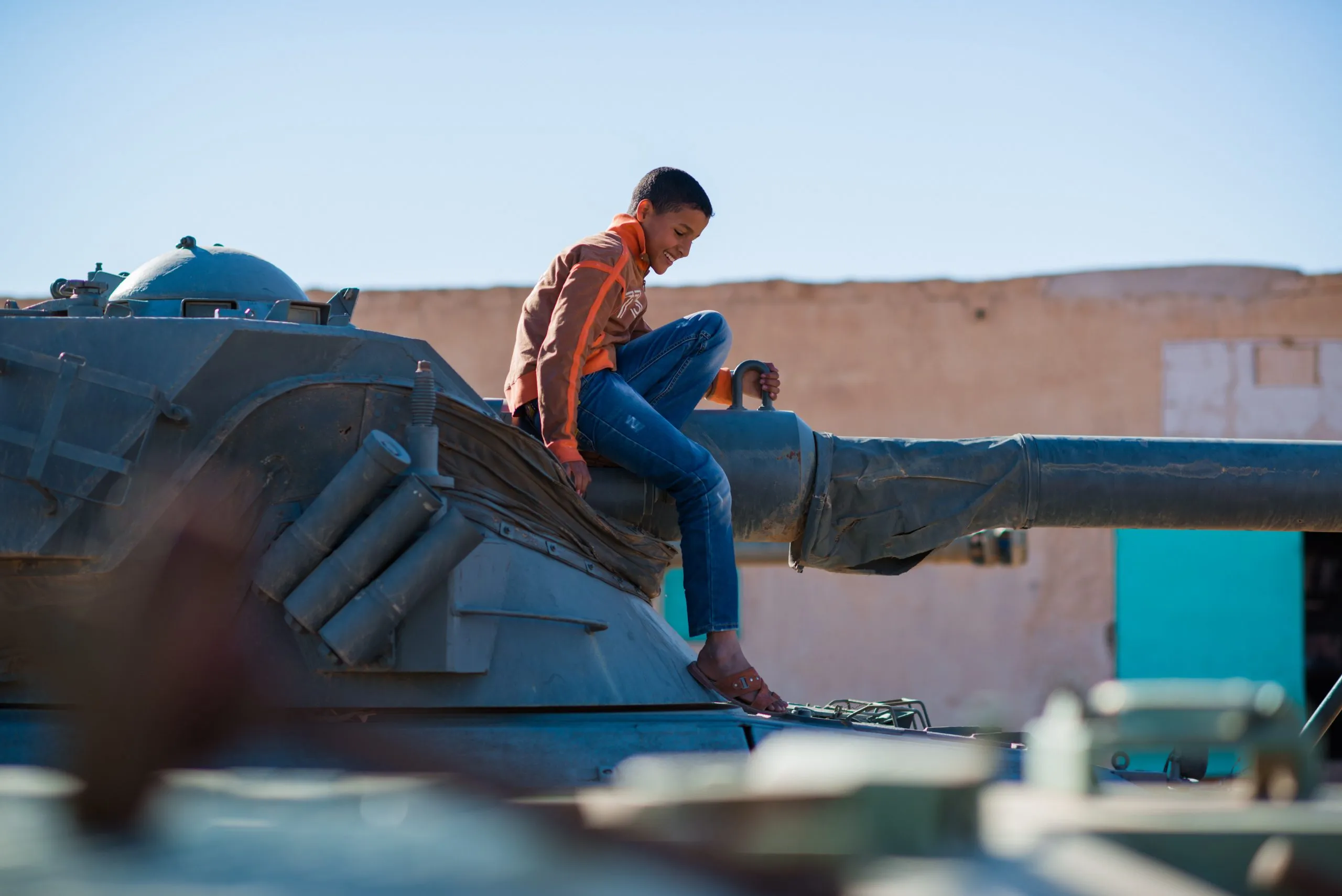 A boy sits atop the gun barrel of an old armoured tank parked under a bright desert sky in Tindouf, Algeria.