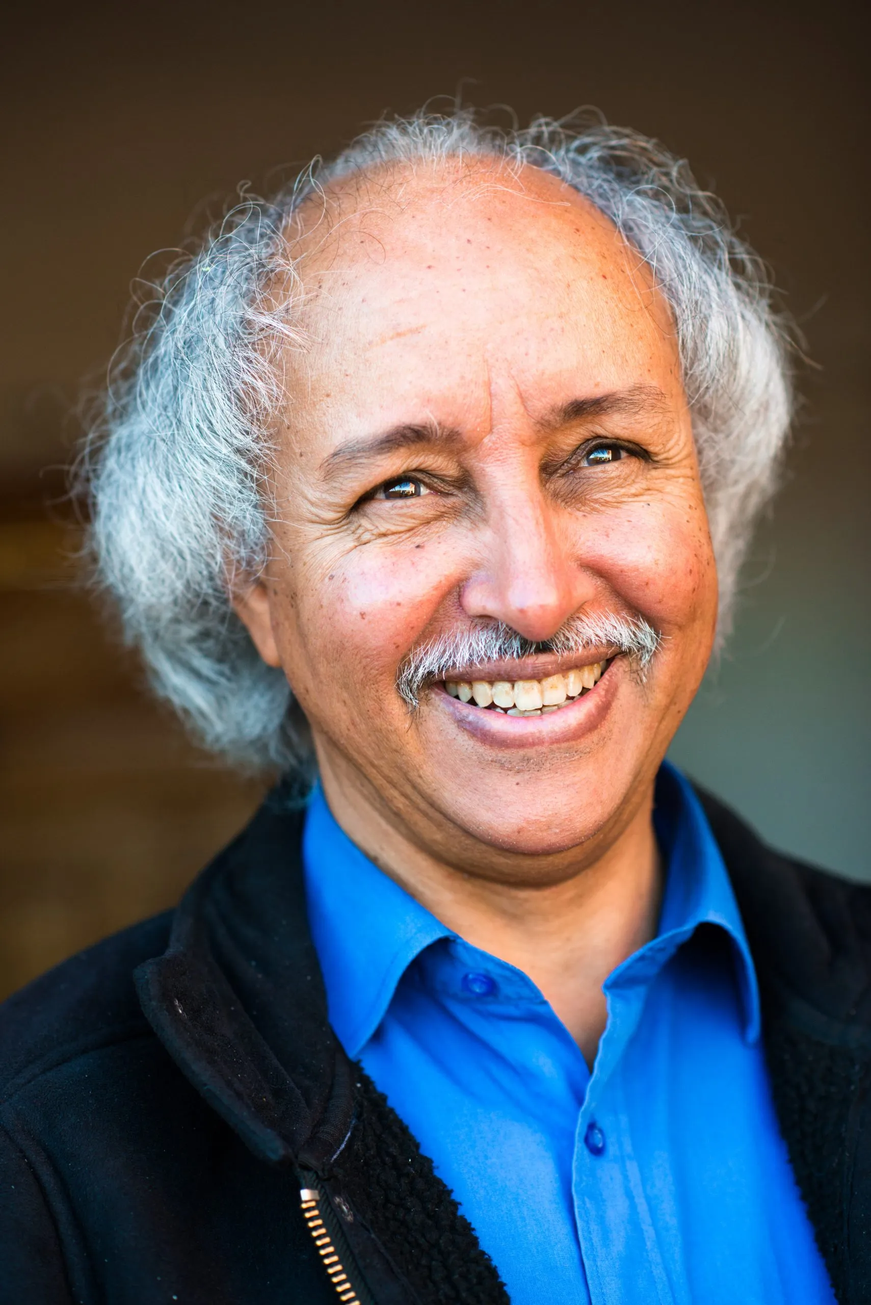 A smiling portrait of an older man with grey hair and a moustache, in a blue shirt and dark fleece, in Tindouf.