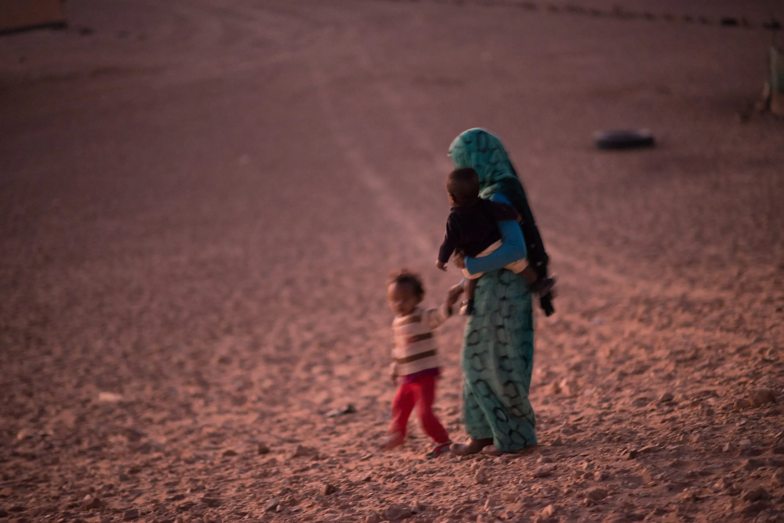 A woman in a patterned dress carries a baby and walks with a small child across the desert at dusk.