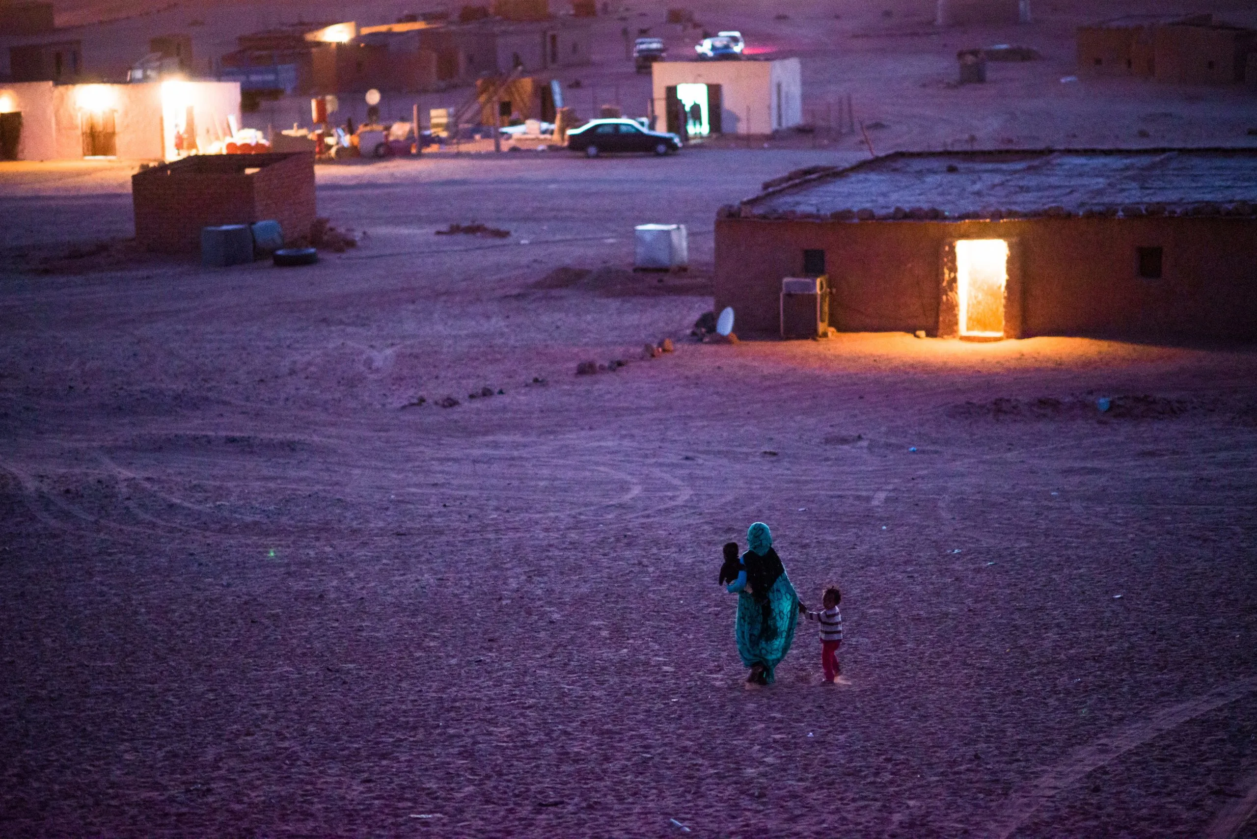 A woman walks two small children home across the open desert at dusk, past low mud-brick houses in the camp.