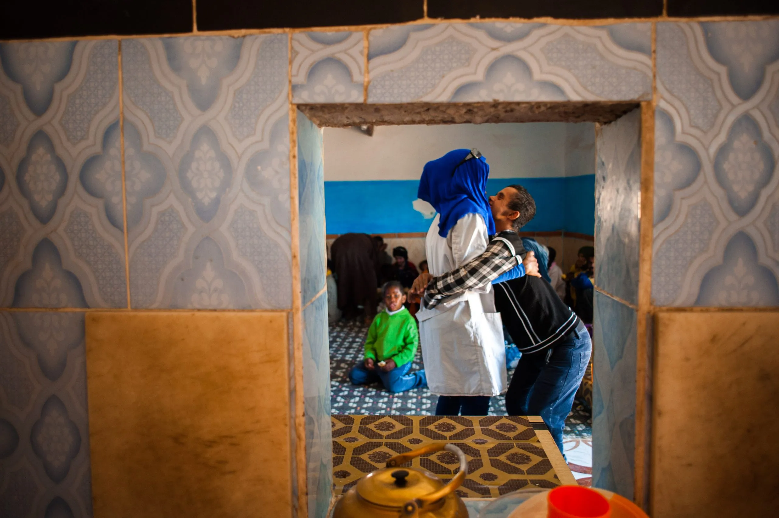 Two women embrace in a tiled doorway as a child looks on from the tea-lit room behind, in a home in Tindouf.