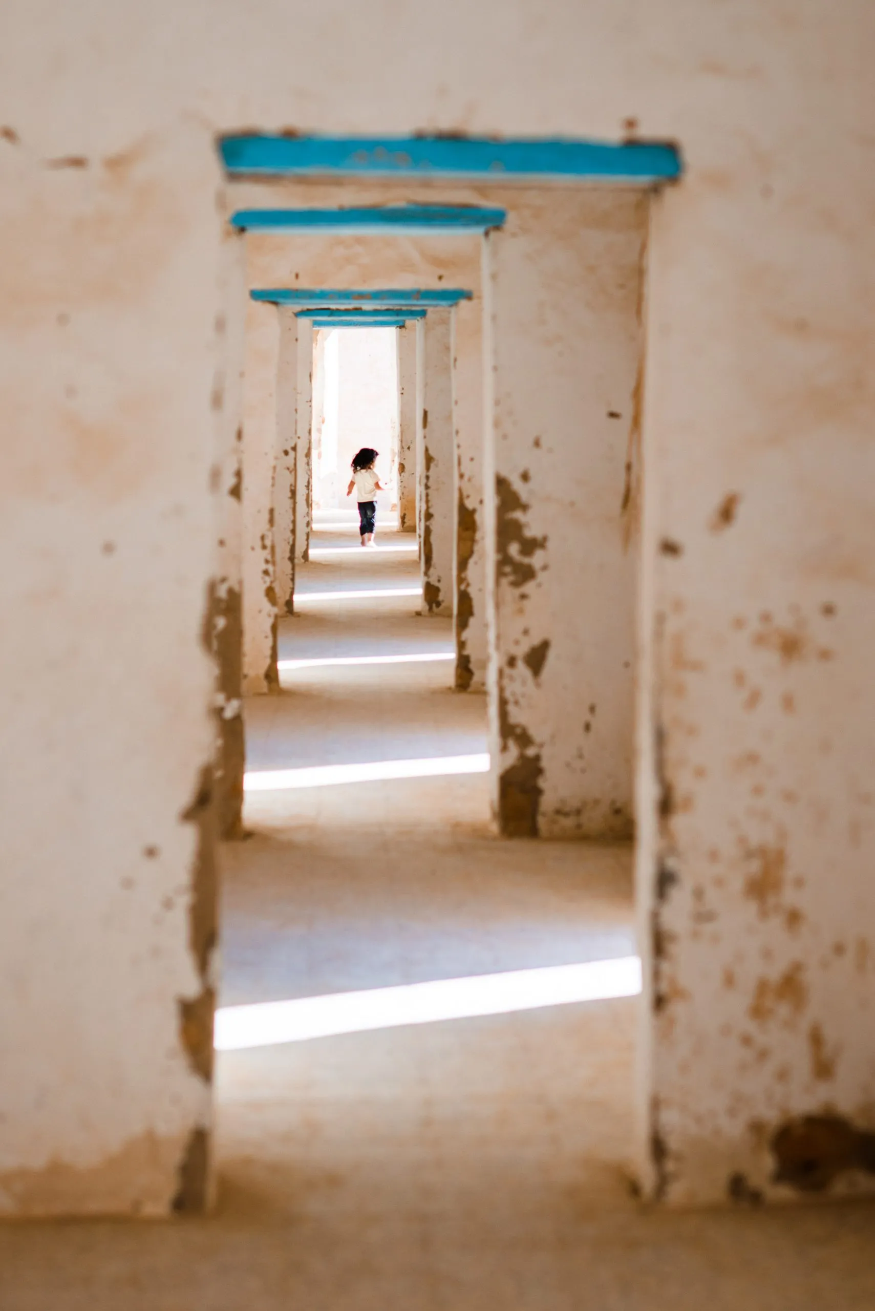 A child runs at the far end of a long corridor of plaster doorways with turquoise lintels, in Tindouf.