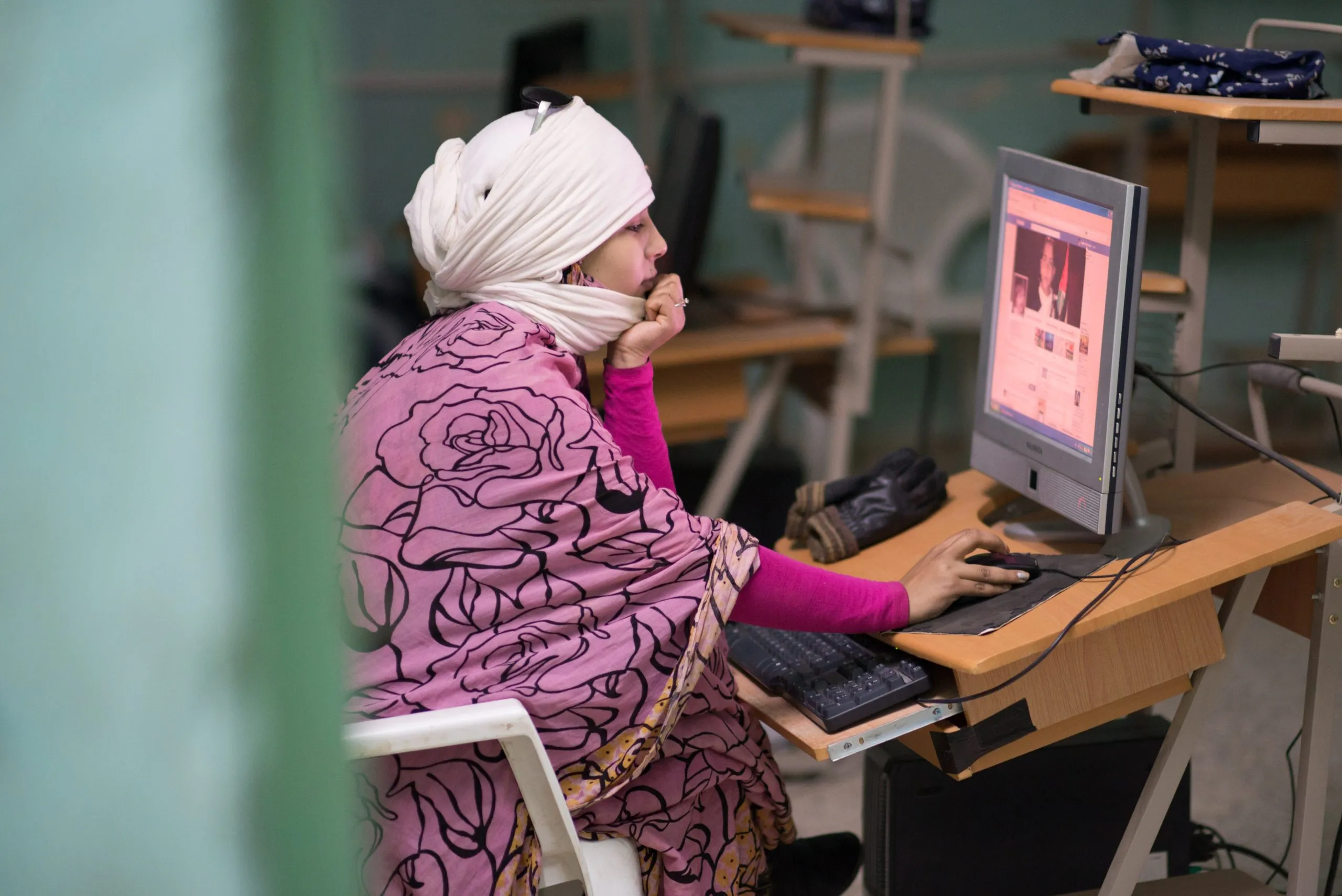 A young woman in a white headscarf and pink top studies at a desktop computer in a classroom in the Tindouf camp.