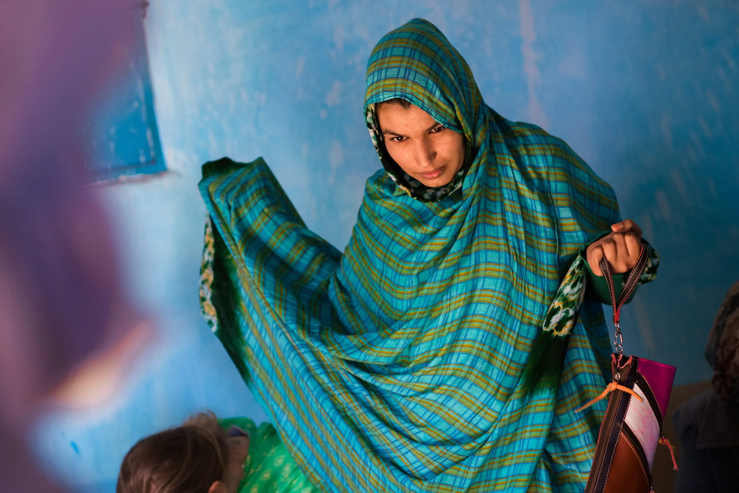 A young woman in a vivid blue-and-green plaid wrap holds a school folder against a blue wall in Tindouf.