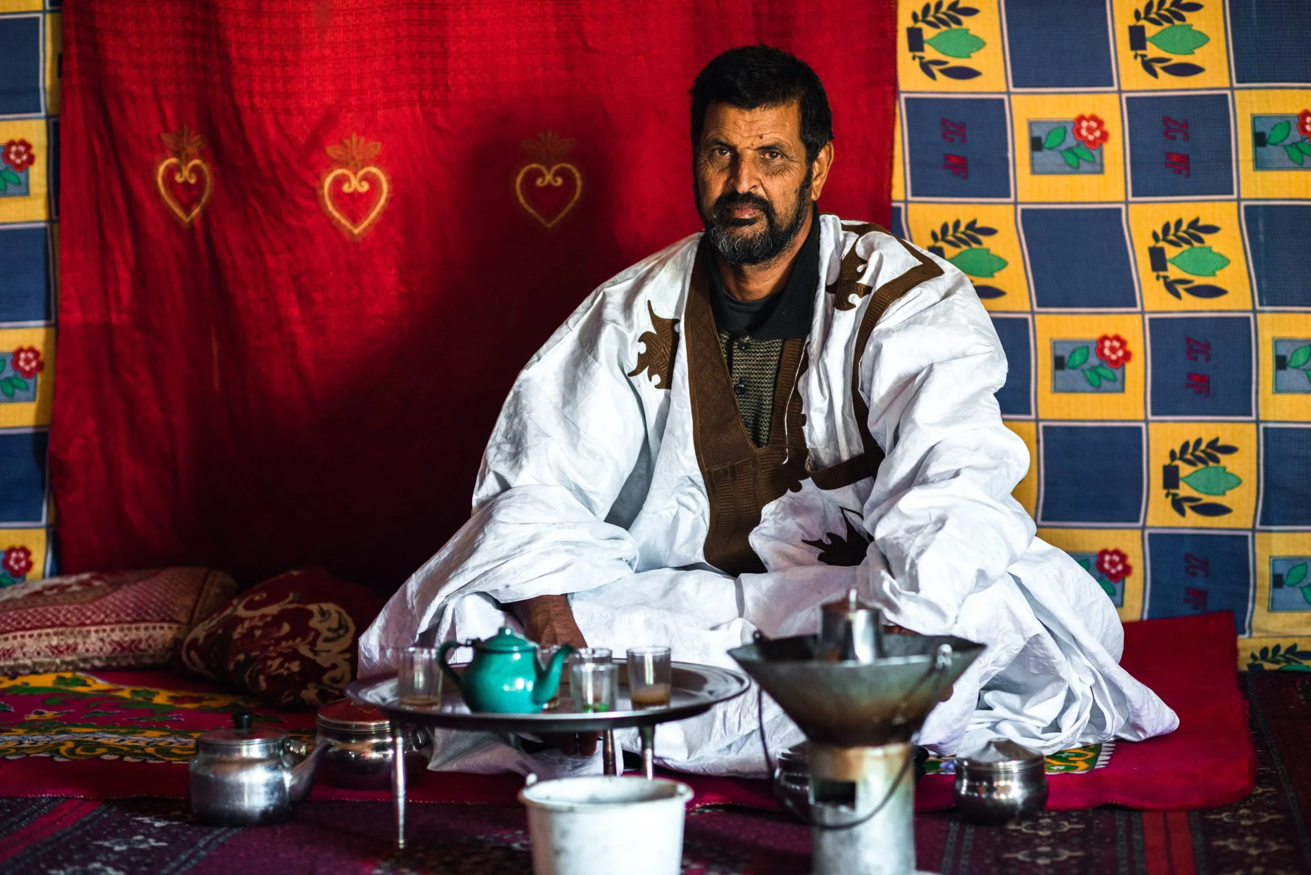 An older man in a white robe and embroidered vest sits with a tea tray on patterned cushions inside a tent.