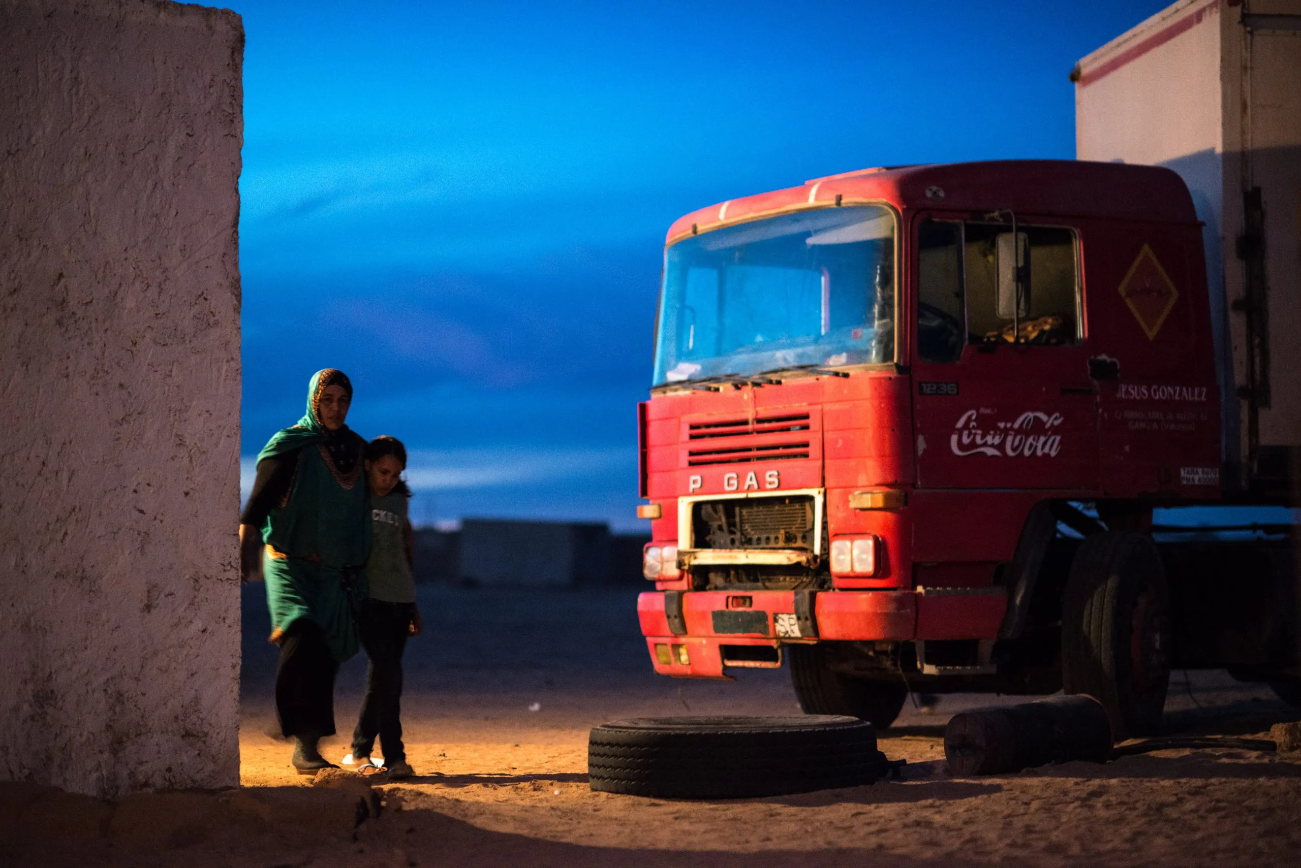 A woman and a small child stand beside a red truck under a deep blue twilight sky, in the camp.