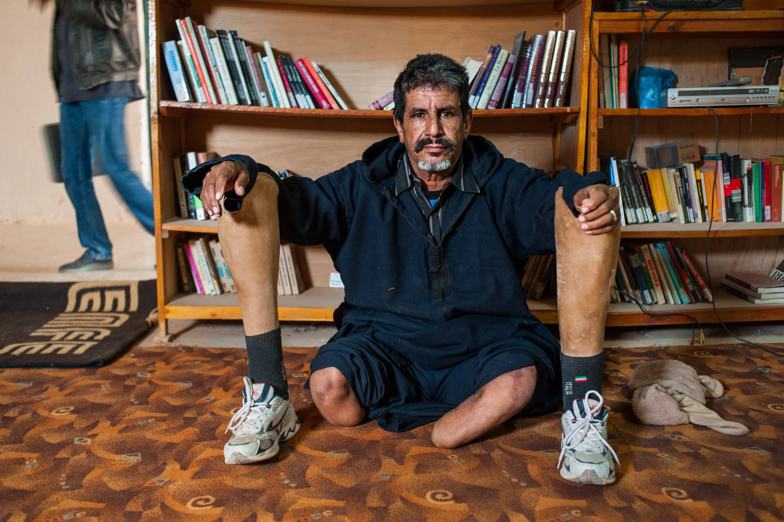 A man with two prosthetic legs sits on a patterned rug in front of bookshelves inside a library in Tindouf.