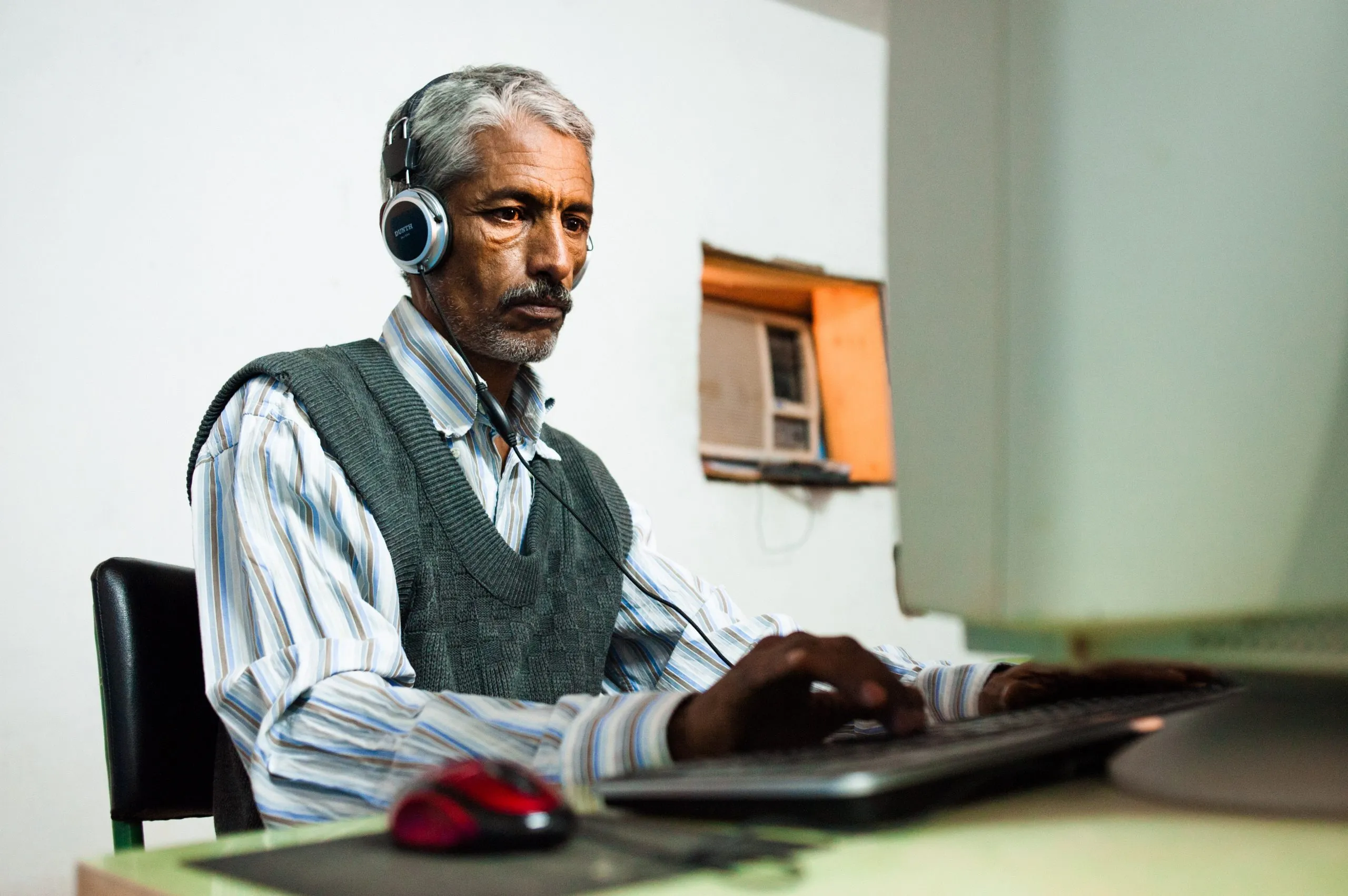 An older man in a striped shirt and grey vest works at a desktop computer wearing headphones, in Tindouf.