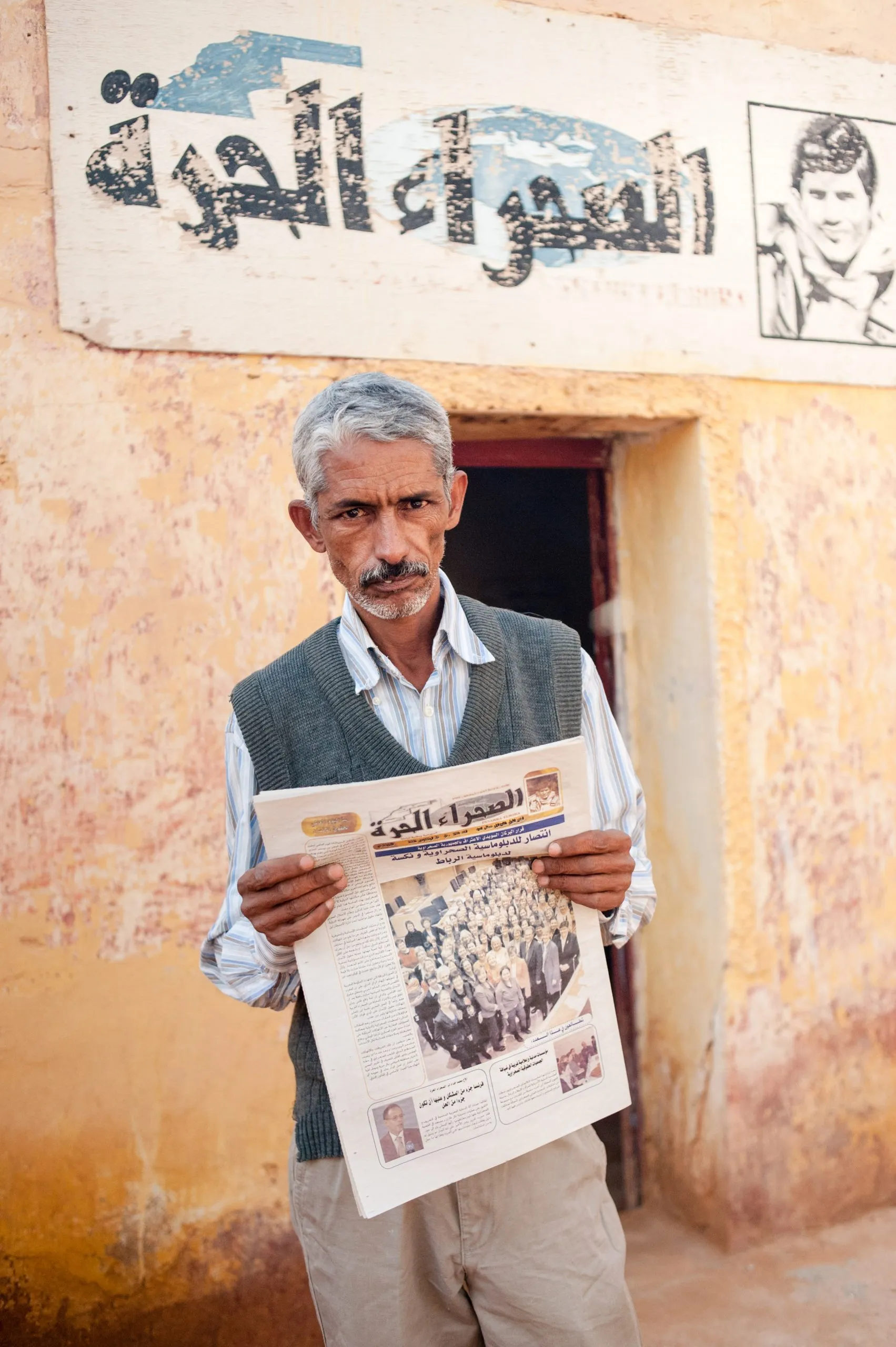 A man holds a copy of the Sahrawi newspaper outside a building with Arabic signage in the Tindouf camps.
