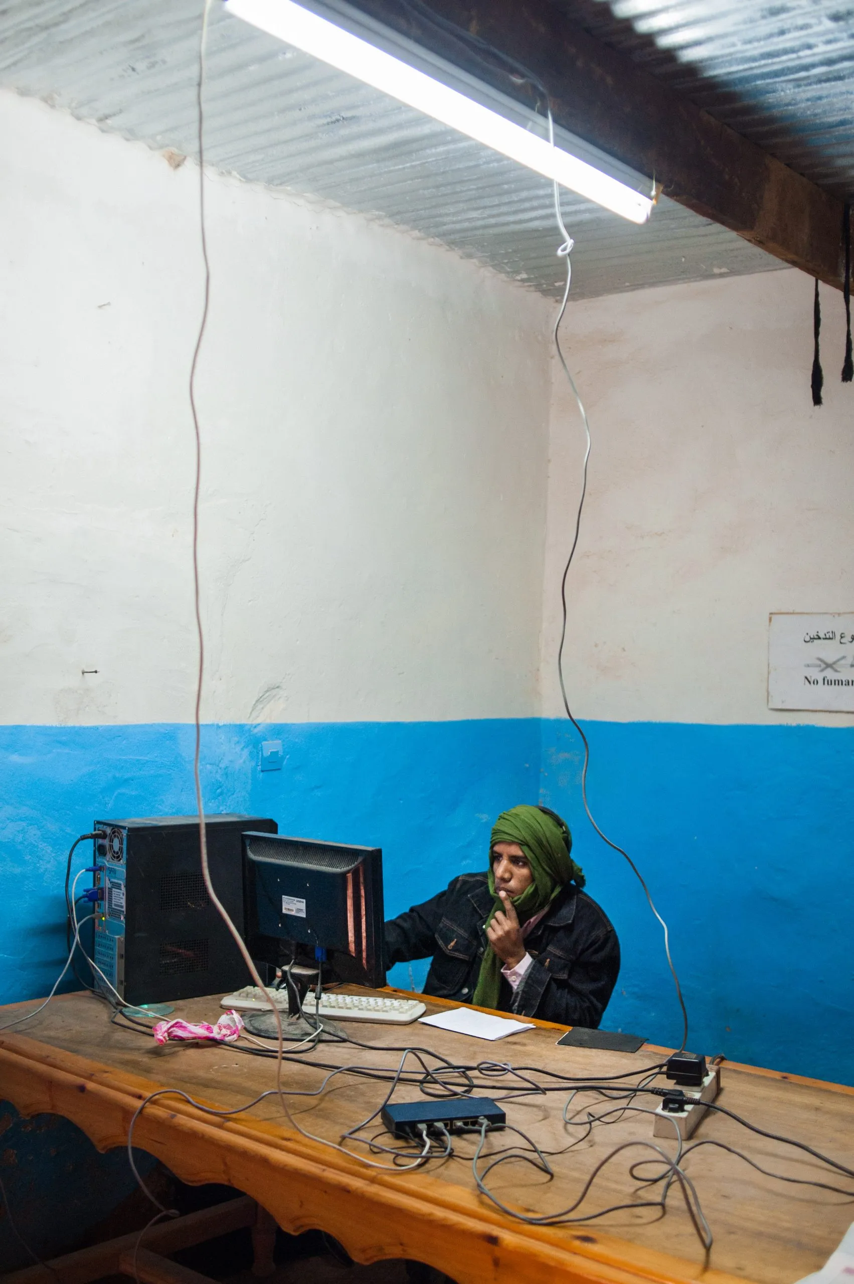 A man with a green scarf around his head works at a computer in a small blue-walled office in Tindouf.