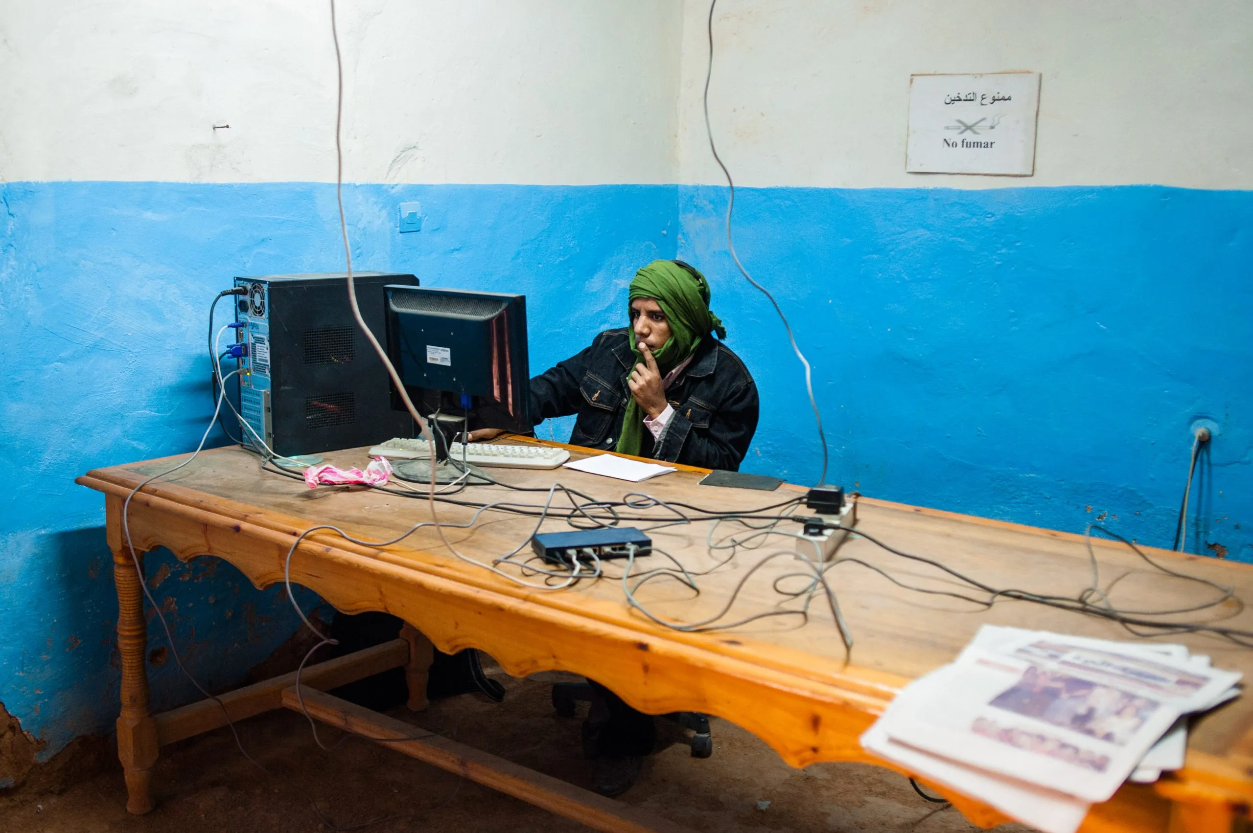 A man in a green head wrap uses a computer at a wooden desk in a blue-and-white striped room, in the Tindouf camp.