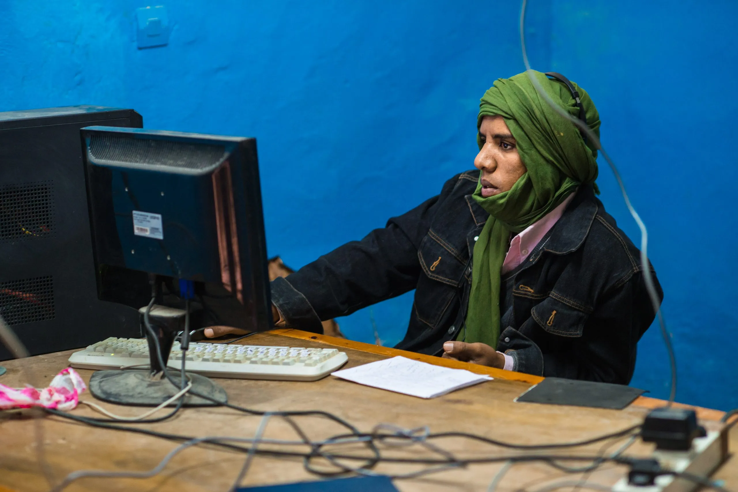 A young person in a green head wrap works at a computer against a vivid blue wall in the Tindouf camp.