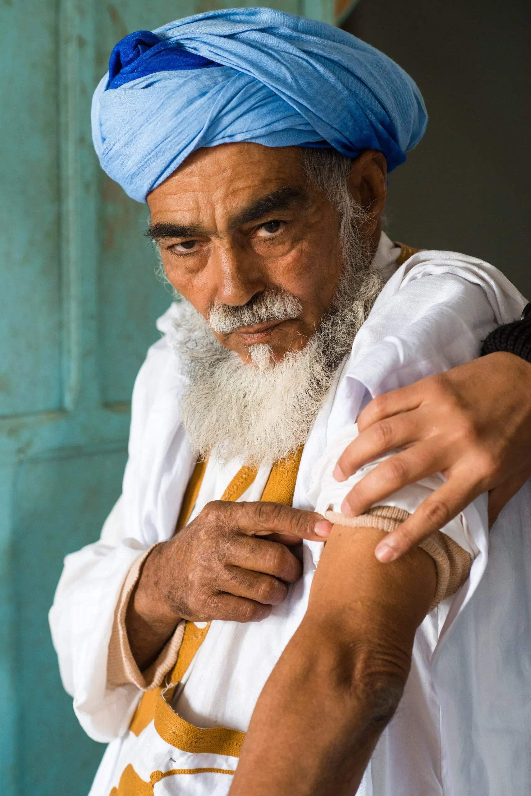 An older man in a blue turban inspects a bandaged arm beside a turquoise door in the Tindouf camp.