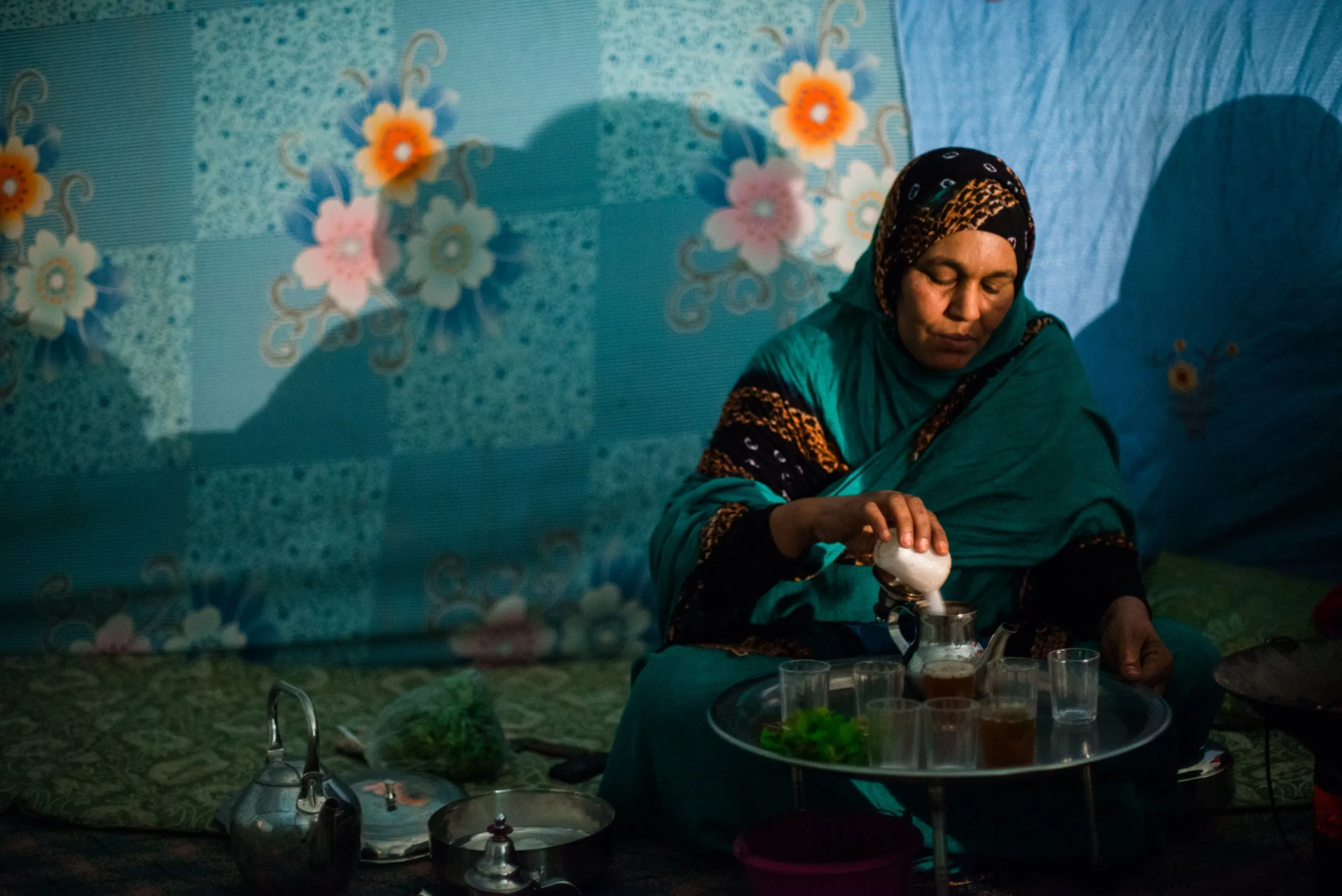 A woman in a turquoise dress and patterned headscarf pours mint tea inside a tent in the Tindouf camps.