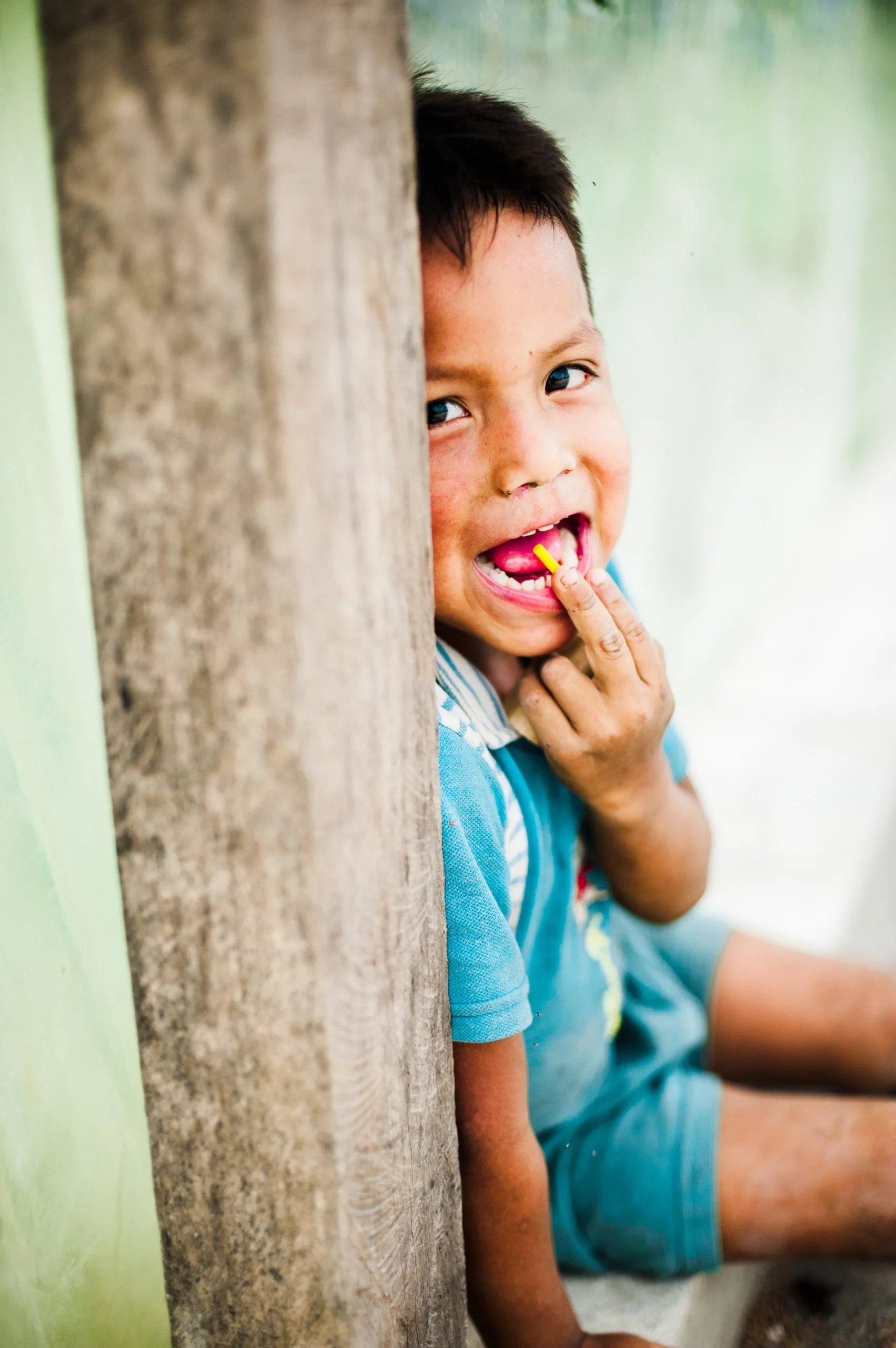 A smiling Ecuadorian boy in a blue shirt peeks from behind a wooden post, showing a small candy held in his teeth.