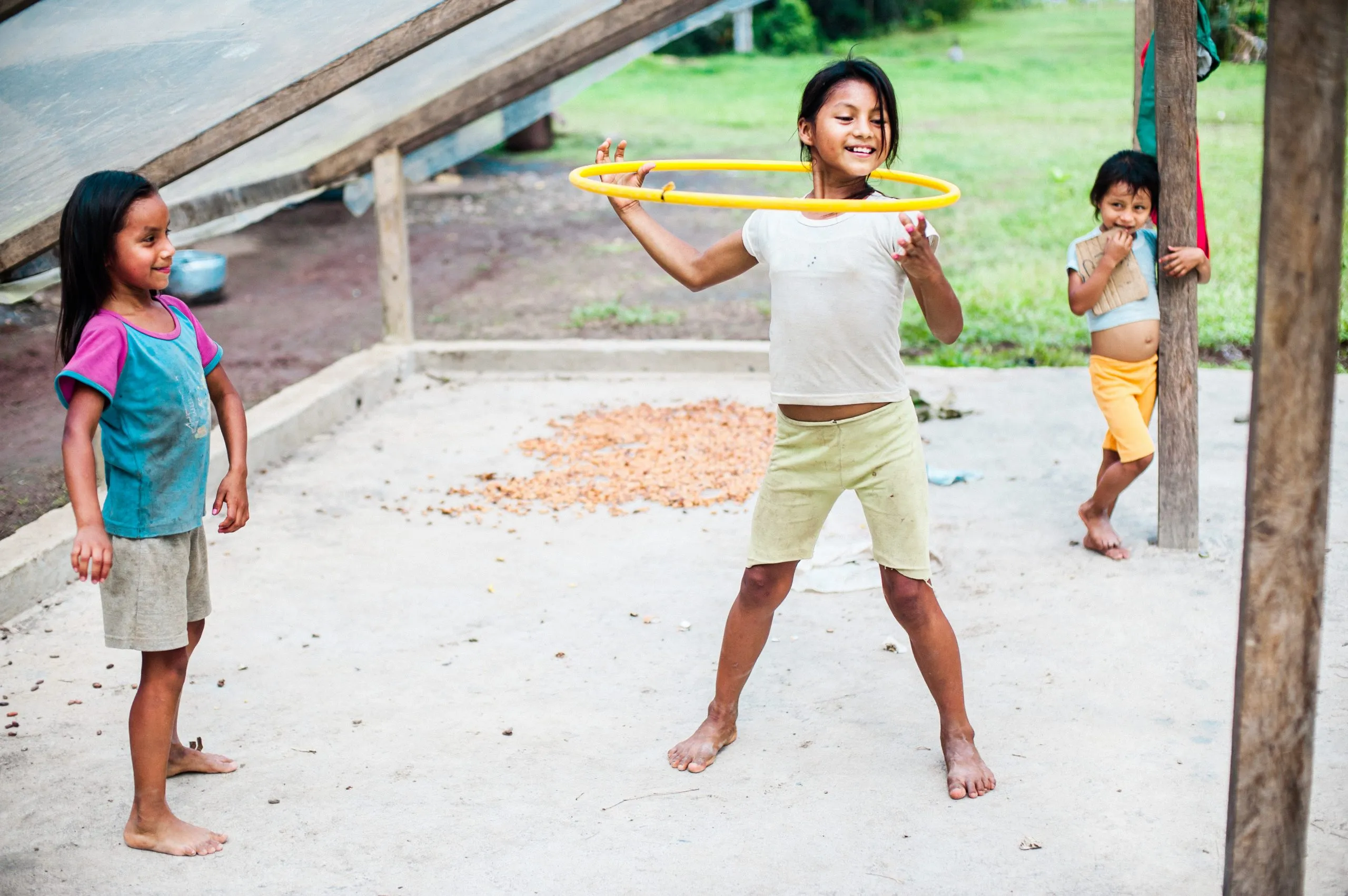 Three Kichwa children play with a yellow plastic hula hoop on the swept earth in front of a palm-thatched hut in the Ecuadorian Amazon.