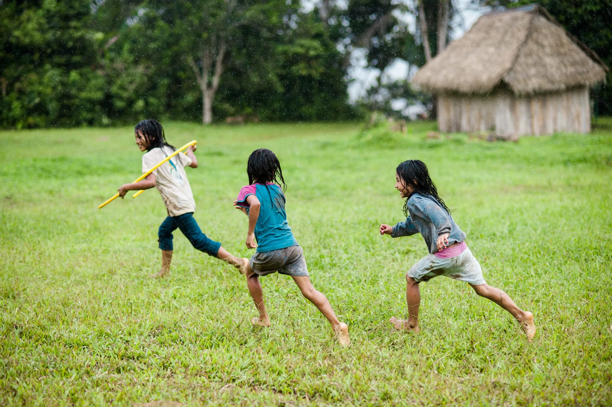 Three Kichwa children run barefoot through grass past a thatched palm hut in an Ecuadorian Amazon village.