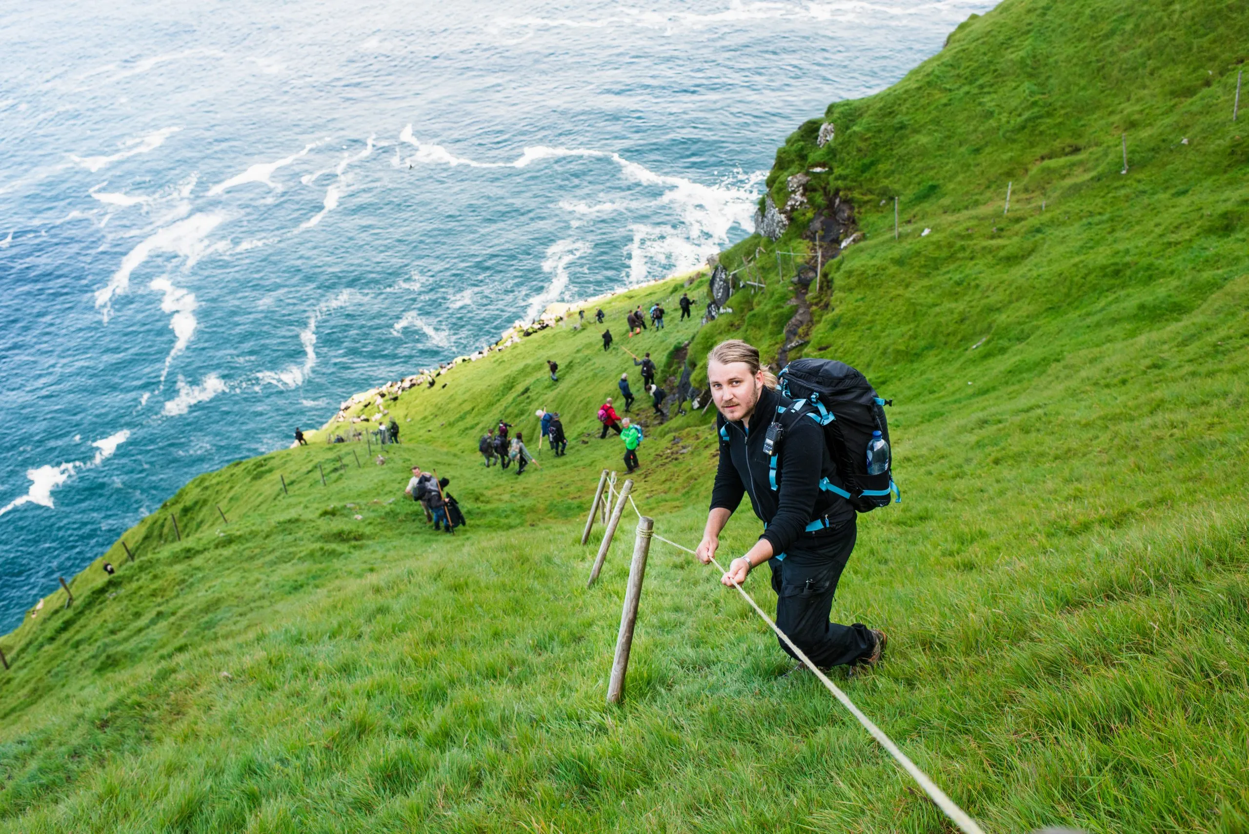 Hikers descend a steep grass slope along a rope handrail above the Atlantic on the Faroese island of Mykines.