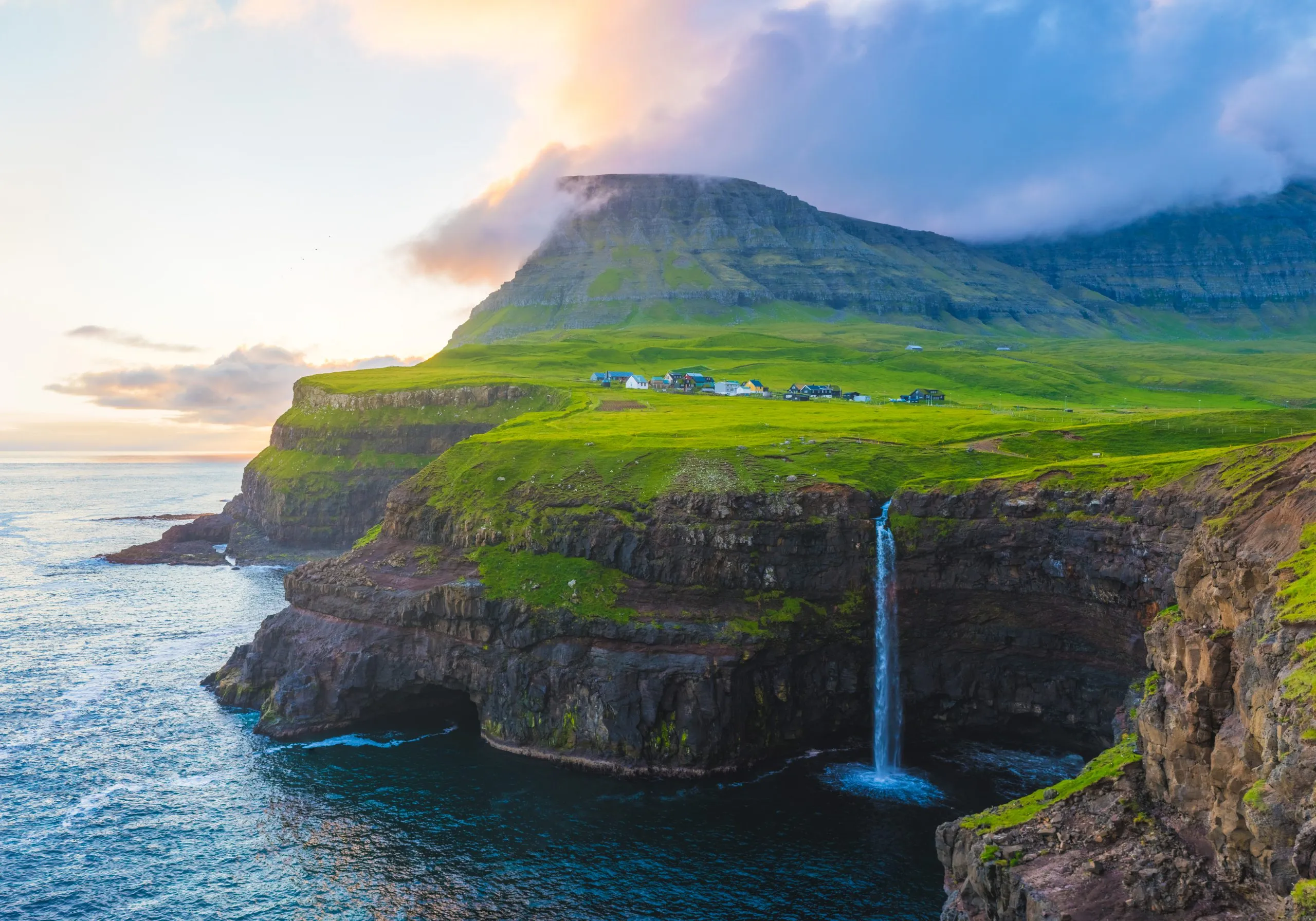Múlafossur waterfall pours from a green Faroese clifftop straight into the sea at sunset, the village of Gásadalur scattered above it.