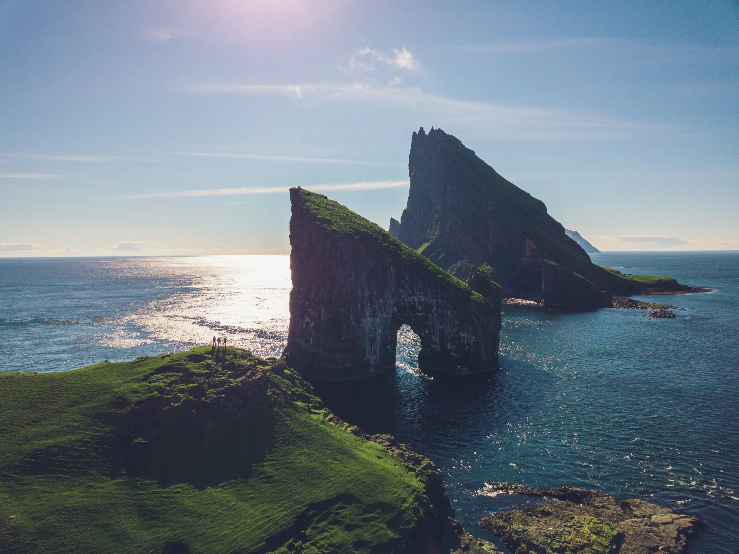 Drone view of the Drangarnir sea-stack arch in the Faroe Islands, three tiny figures standing on the green clifftop in the foreground.