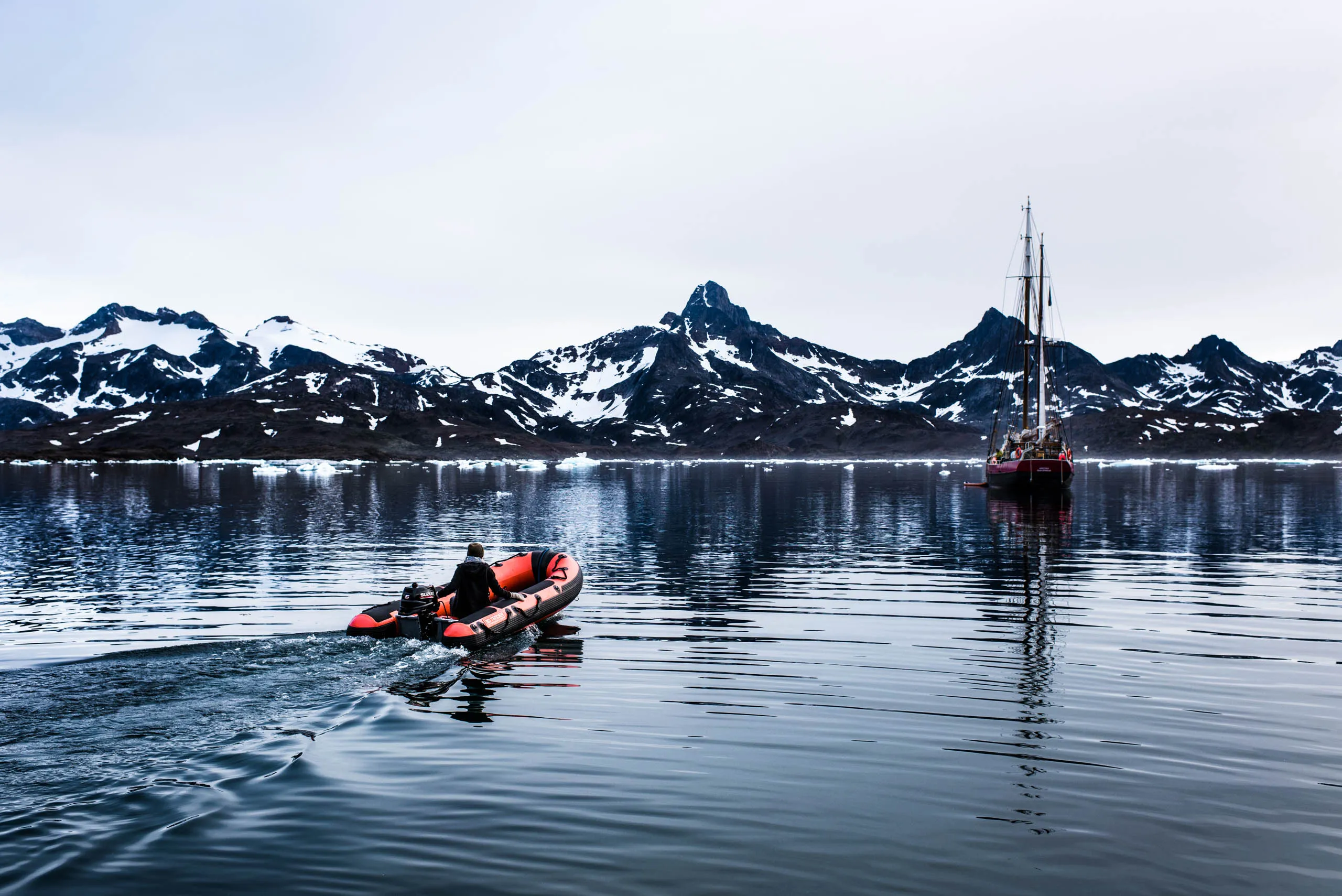 Red inflatable dinghy motoring toward a tall-masted sailboat on a glassy Arctic fjord