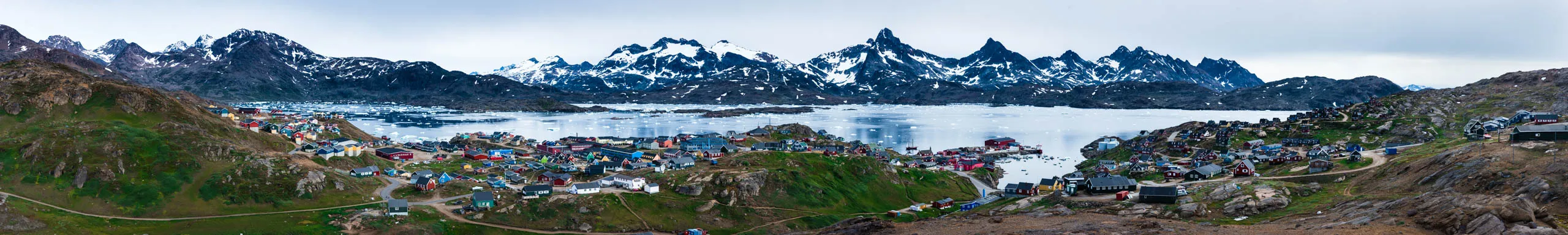 Wide panorama of a Greenlandic town ringed by jagged snow-capped peaks and a frozen inlet