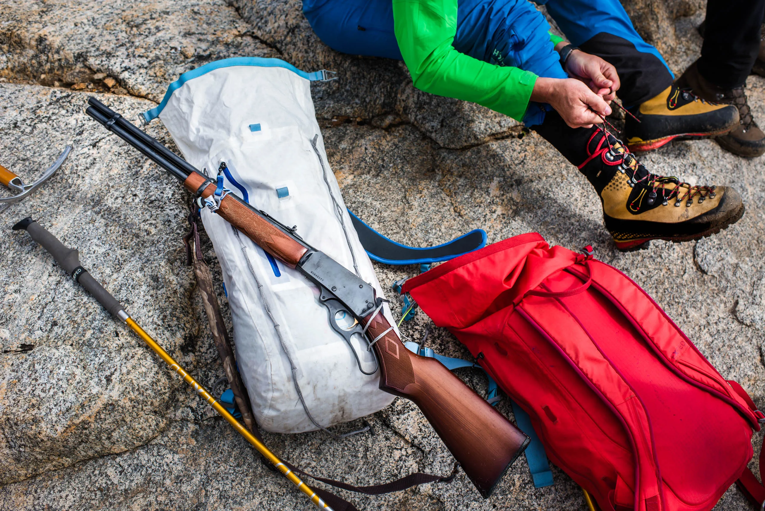 Hiker lacing boots beside a rifle and ice axe on granite rocks in the Arctic