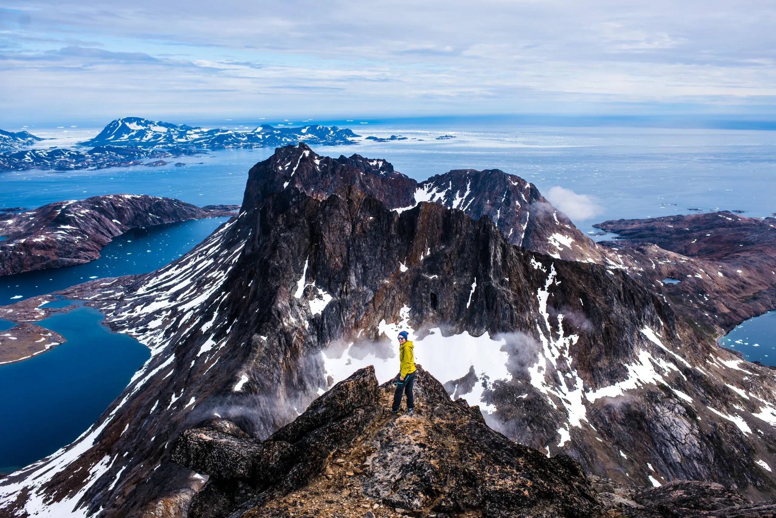 A person in a yellow jacket stands on a rocky summit overlooking a vast iceberg-strewn fjord and snow-capped mountains in East Greenland.