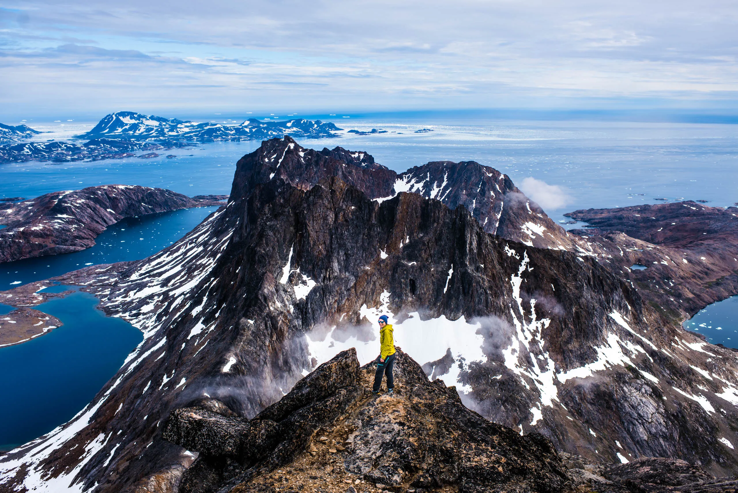 Lone hiker in yellow jacket standing on a ridge above dramatic snow-streaked Greenland peaks and icy fjords