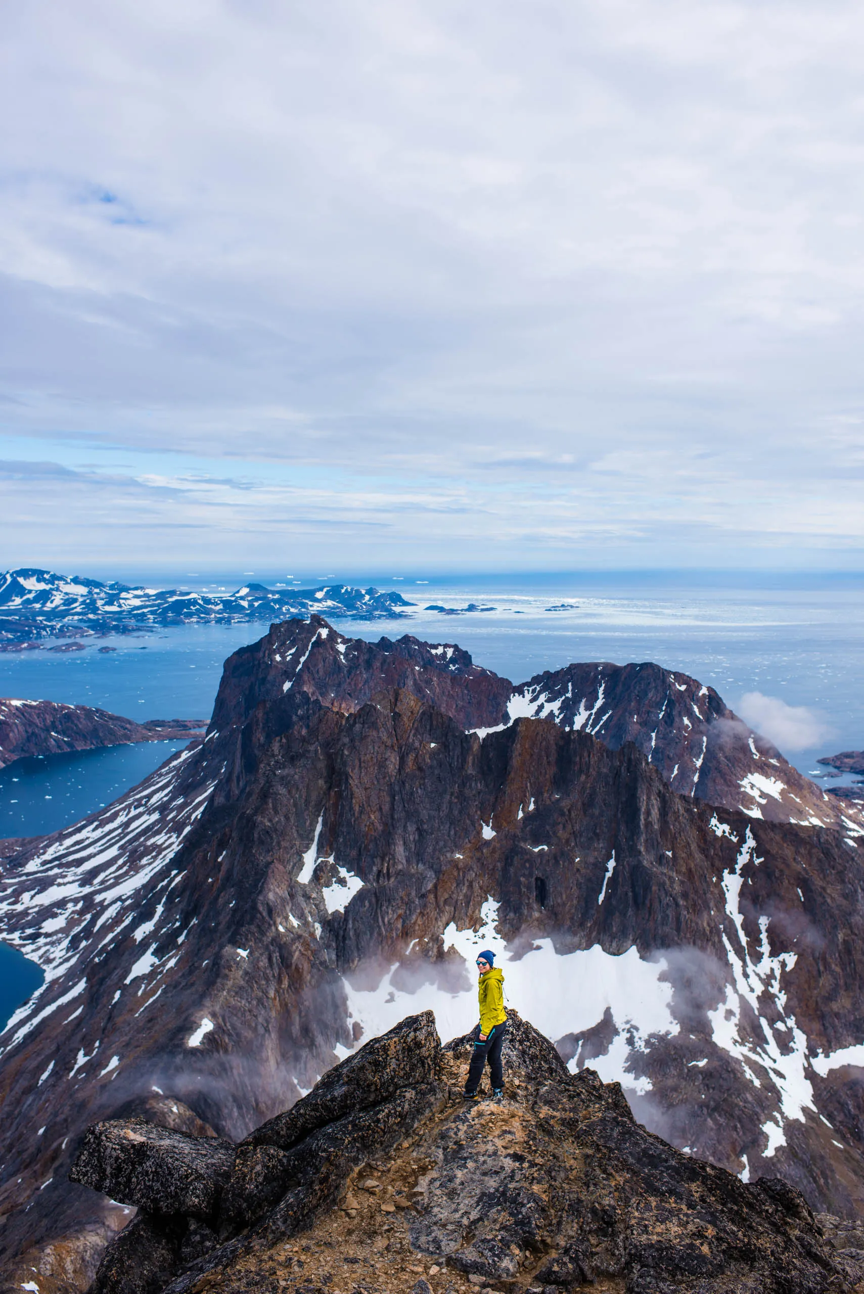 Climber on a rocky summit ridge dwarfed by towering snow-covered peaks above an Arctic sea